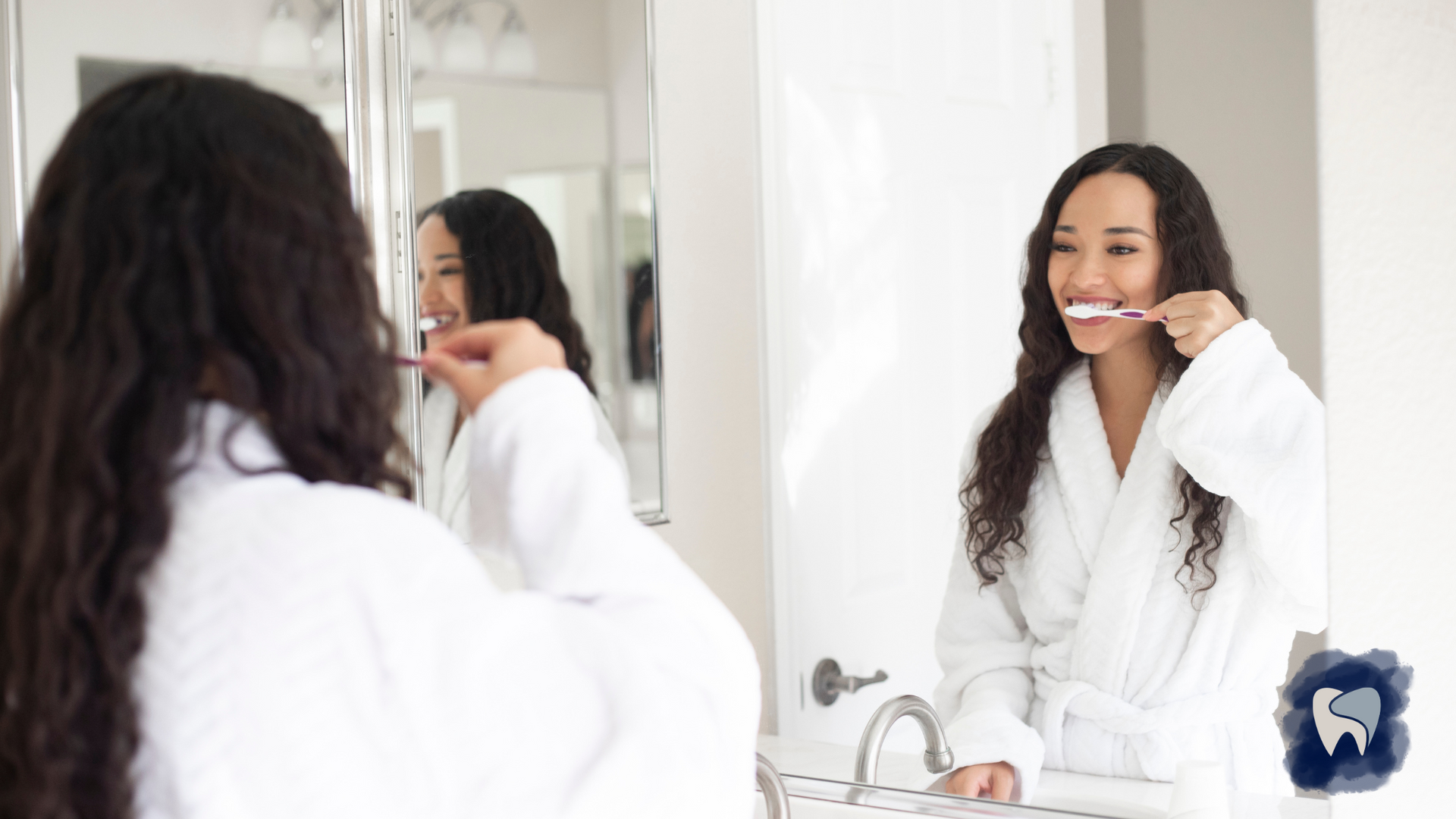 Woman in a white robe brushing her teeth in a bathroom, looking in a mirror, smiling.