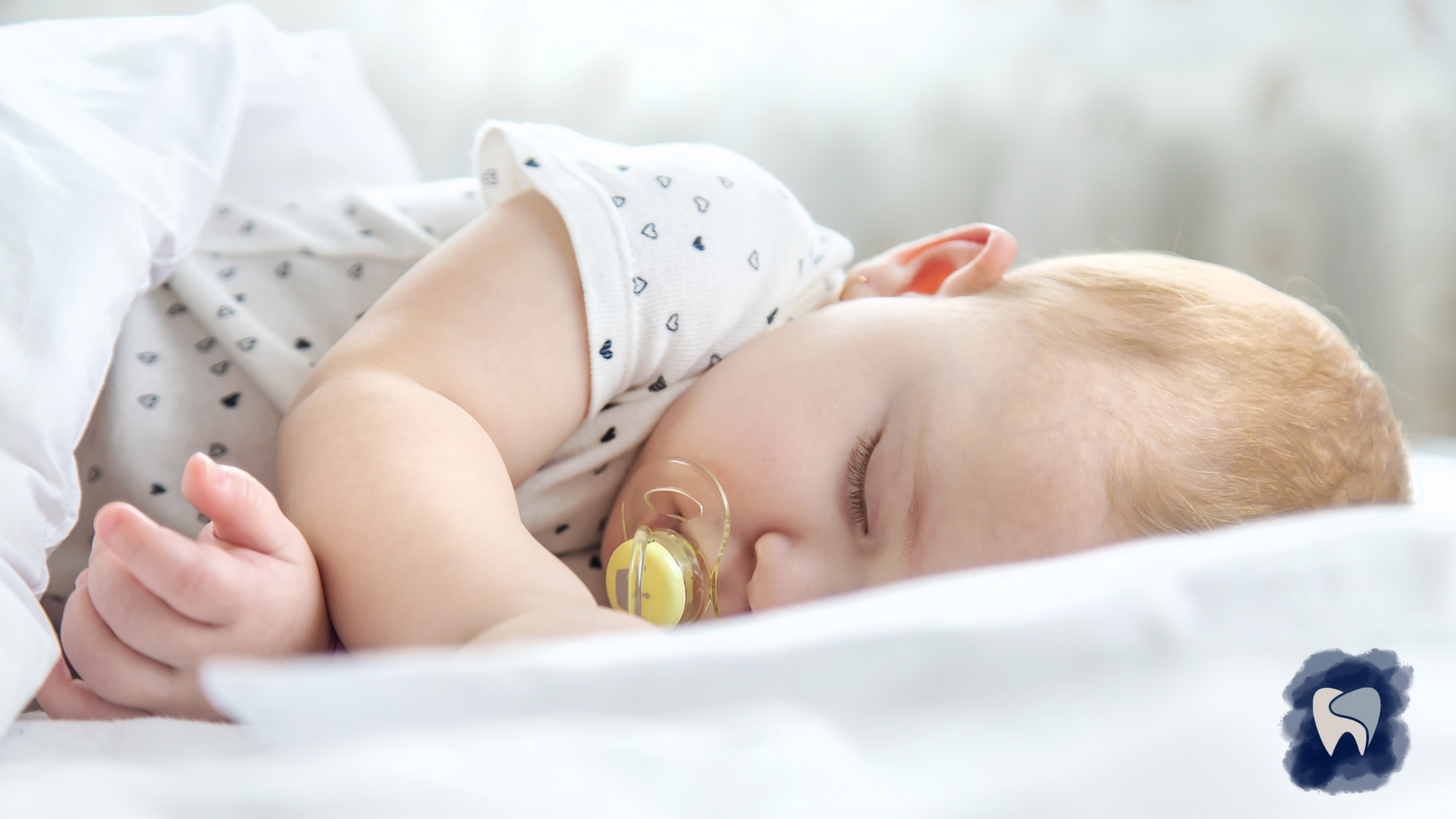 Baby sleeping with a pacifier on a white bed, arm raised.