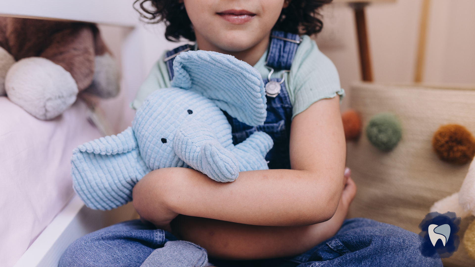 Child hugging a light blue stuffed elephant; wearing denim overalls, seated on the floor.