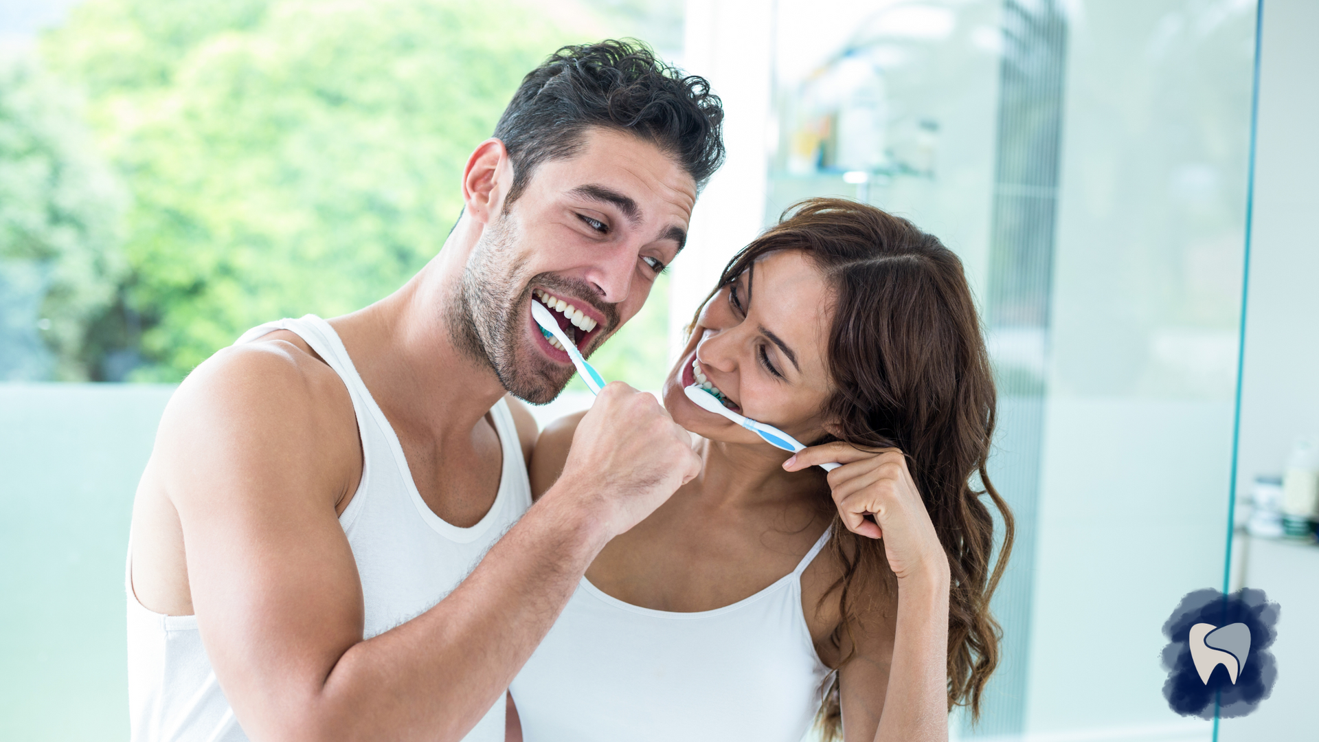 Couple smiling while brushing teeth in a bathroom, near a mirror.