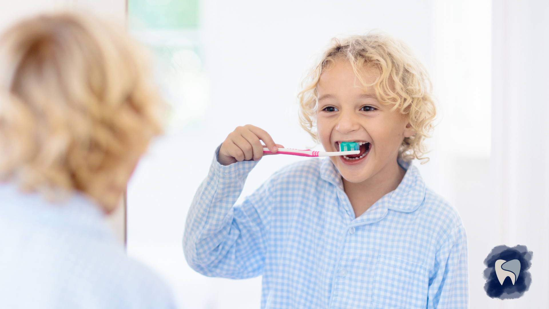 Boy brushing teeth in front of a mirror, smiling, blue pajamas, white bathroom.