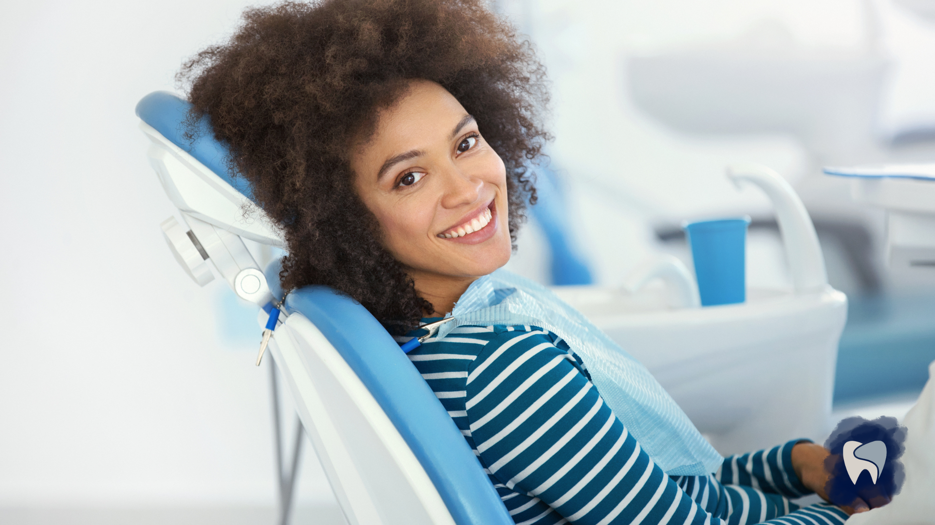 Woman smiling in a dental chair; bright lighting, light blue and white dental equipment visible.