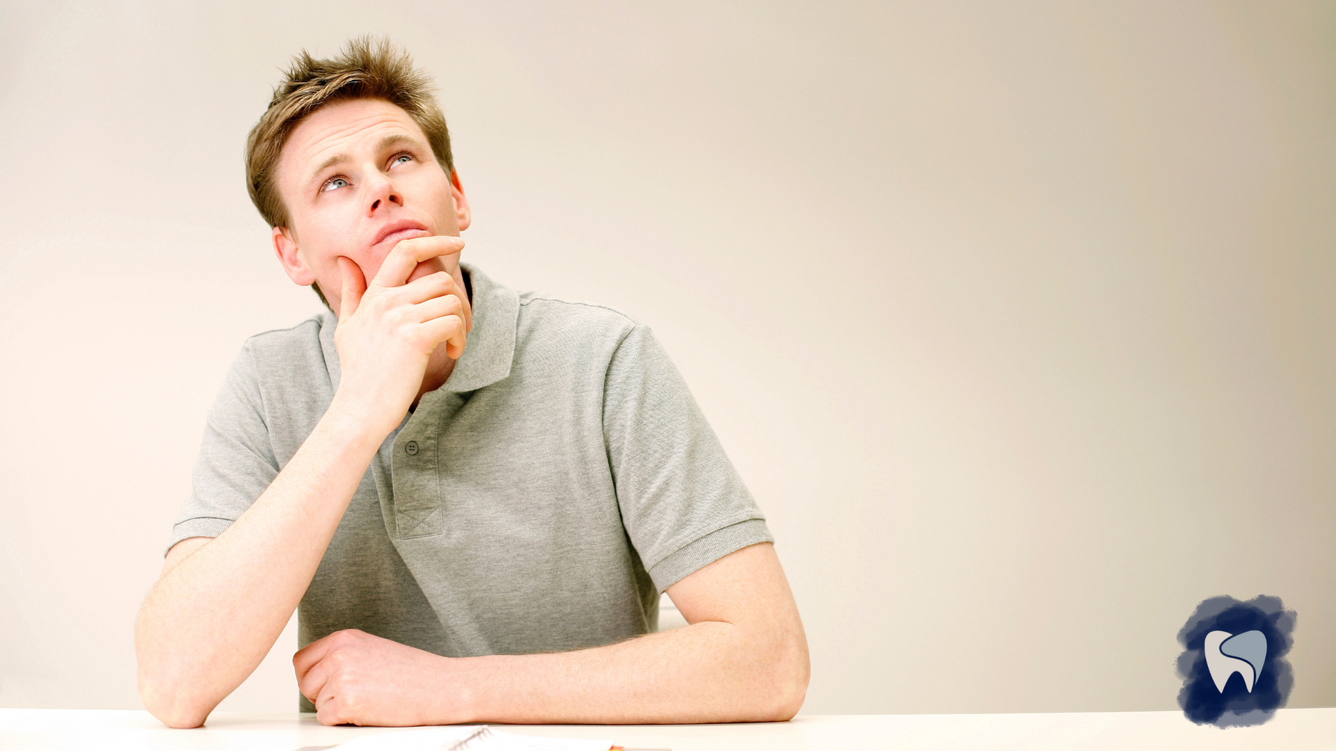 Man in grey shirt, seated, looking upwards with hand on chin, thinking. Light background.