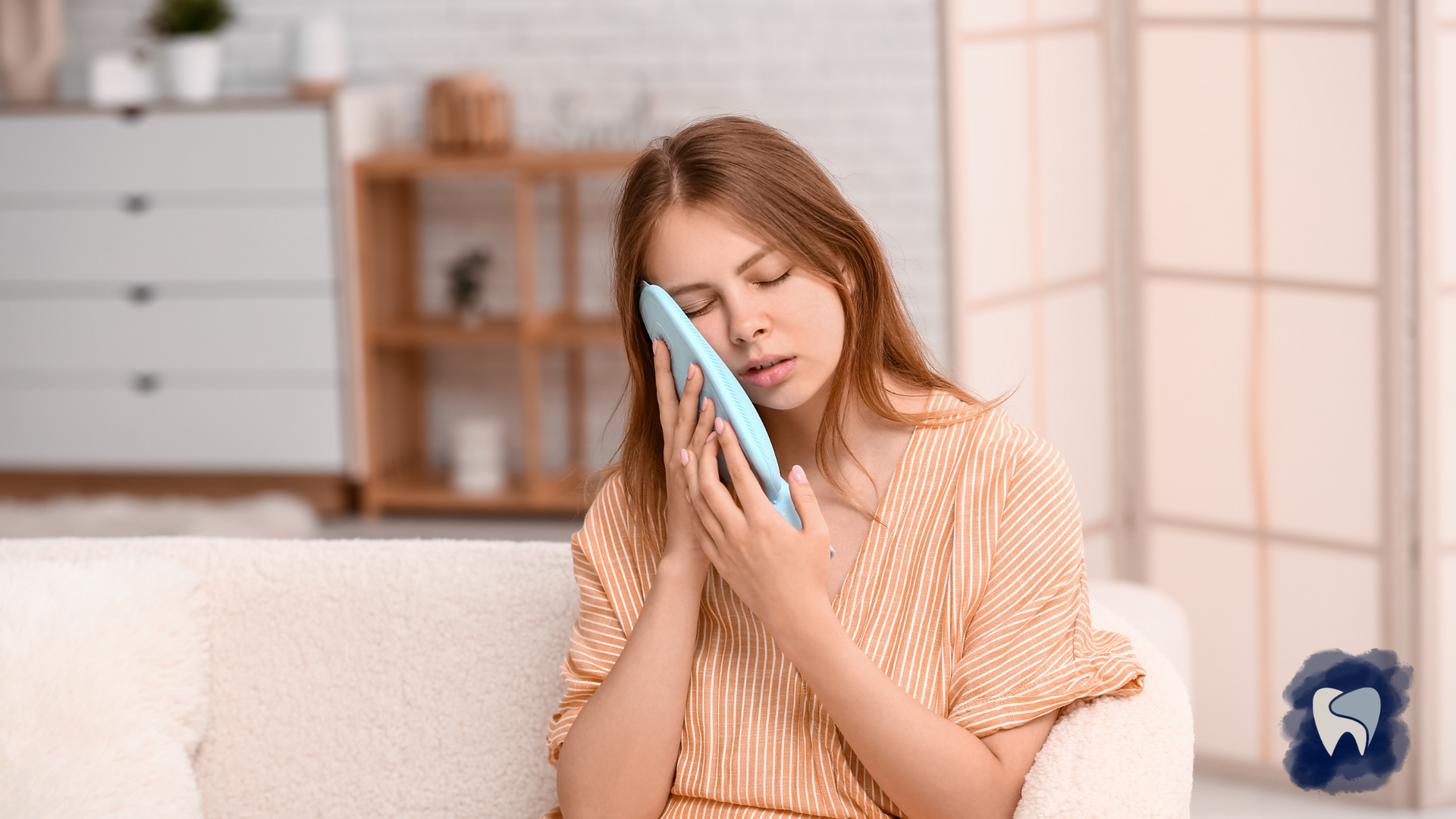 Woman on couch holding ice pack to her cheek, presumably due to toothache; light-filled room.