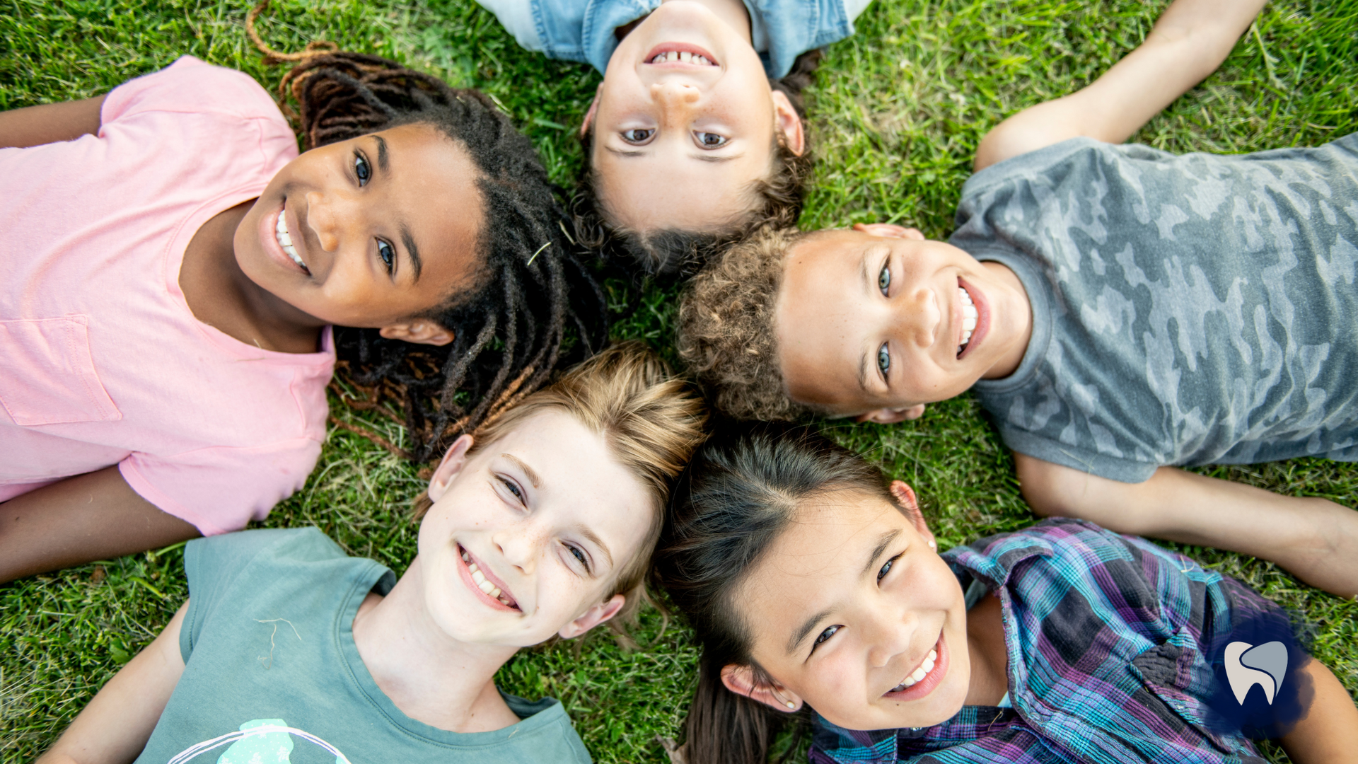 Children lying in a circle on grass, smiling up at the camera.