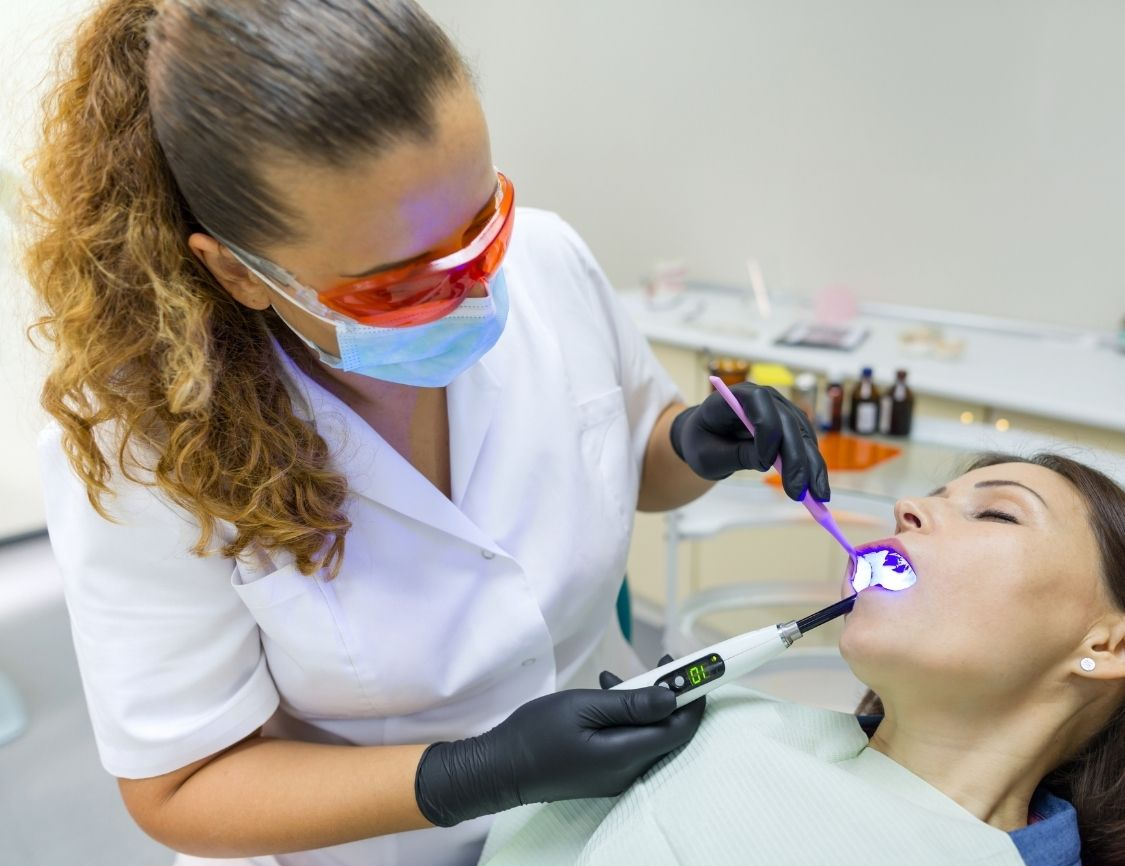 Dentist using a curing light on a patient's tooth. Doctor wearing protective gear, patient in dental chair.