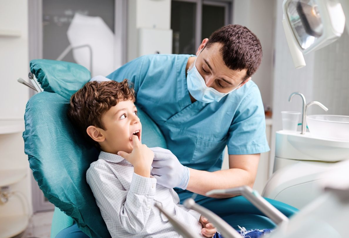 Dentist examining a young boy's mouth in a dental chair. Boy has surprised expression, blue scrubs.