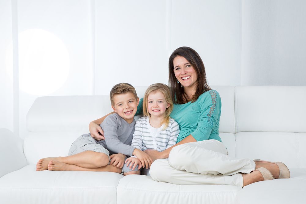 Woman and two smiling children on a white couch. Woman wears teal shirt, kids wear grey and striped shirts. Bright, white room.