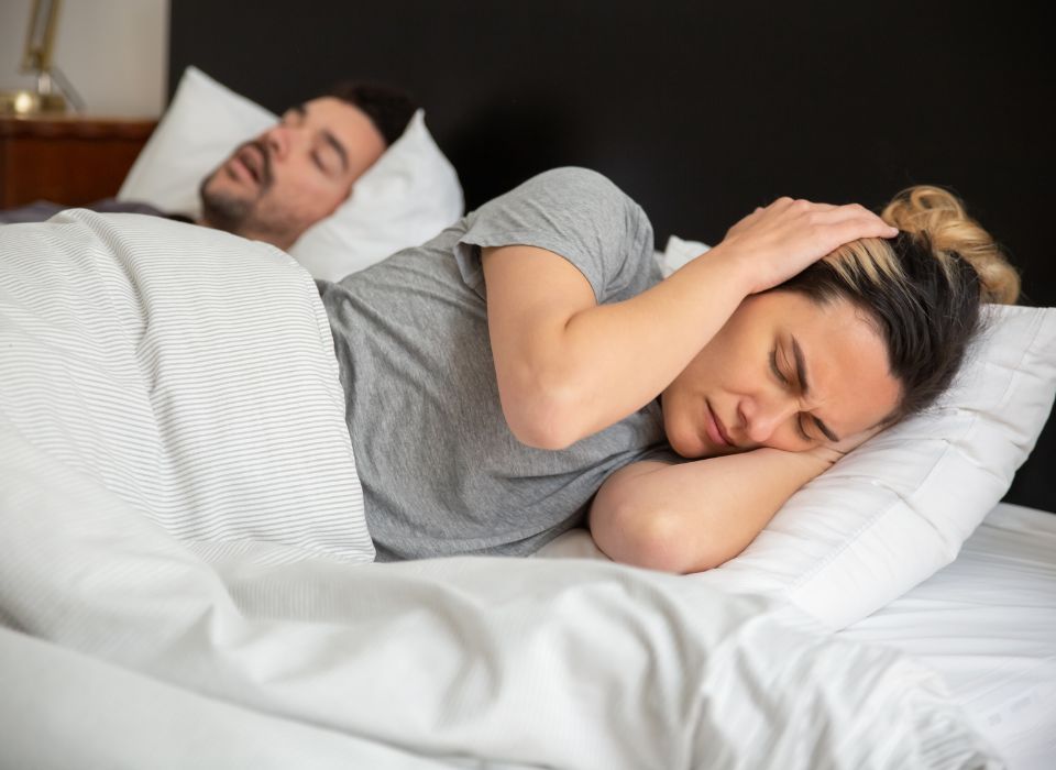 Woman in bed covers ears, frustrated, while man snores beside her. Bedroom setting.