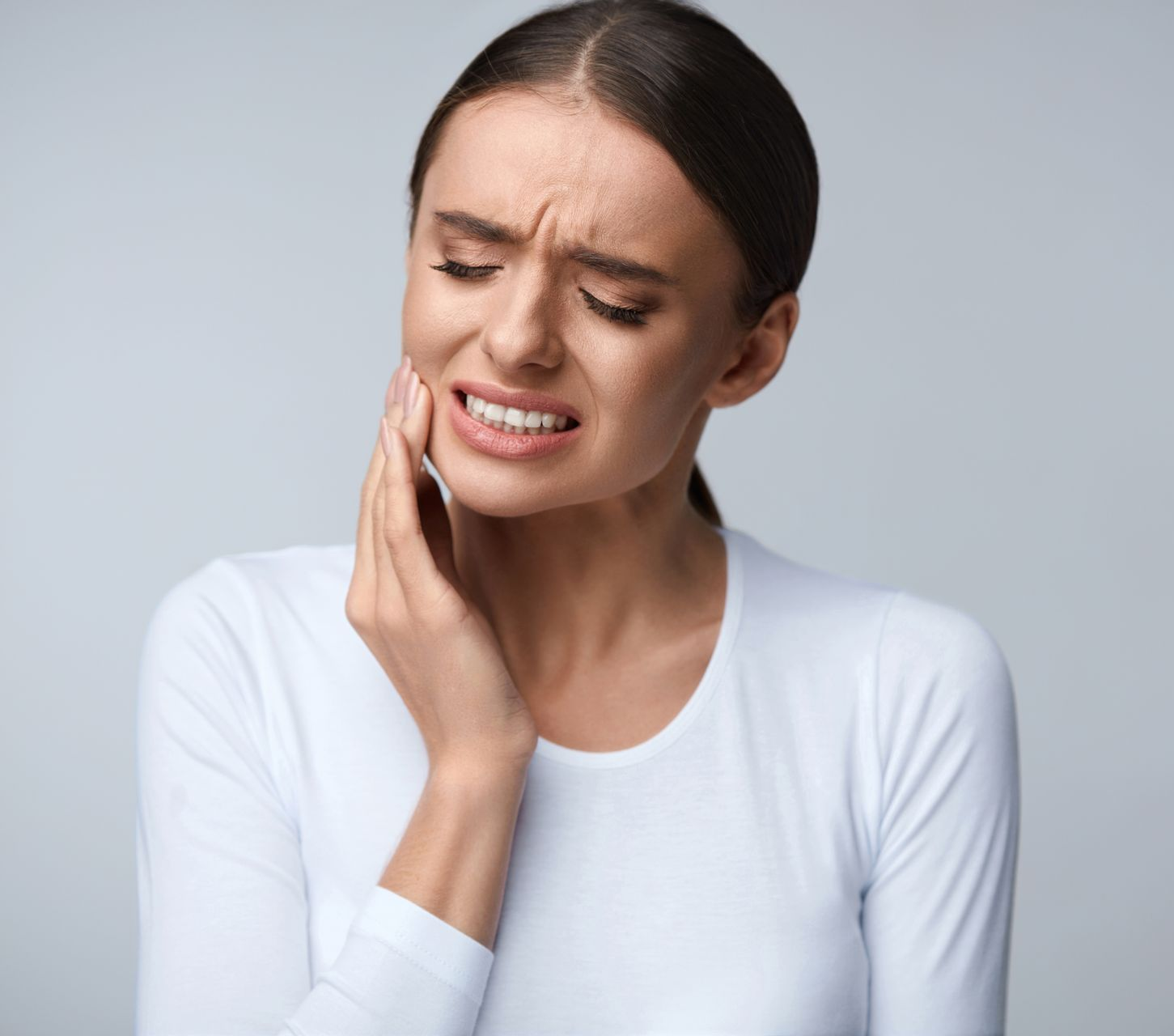 Woman in white shirt with pained expression, hand on cheek, suggesting toothache.