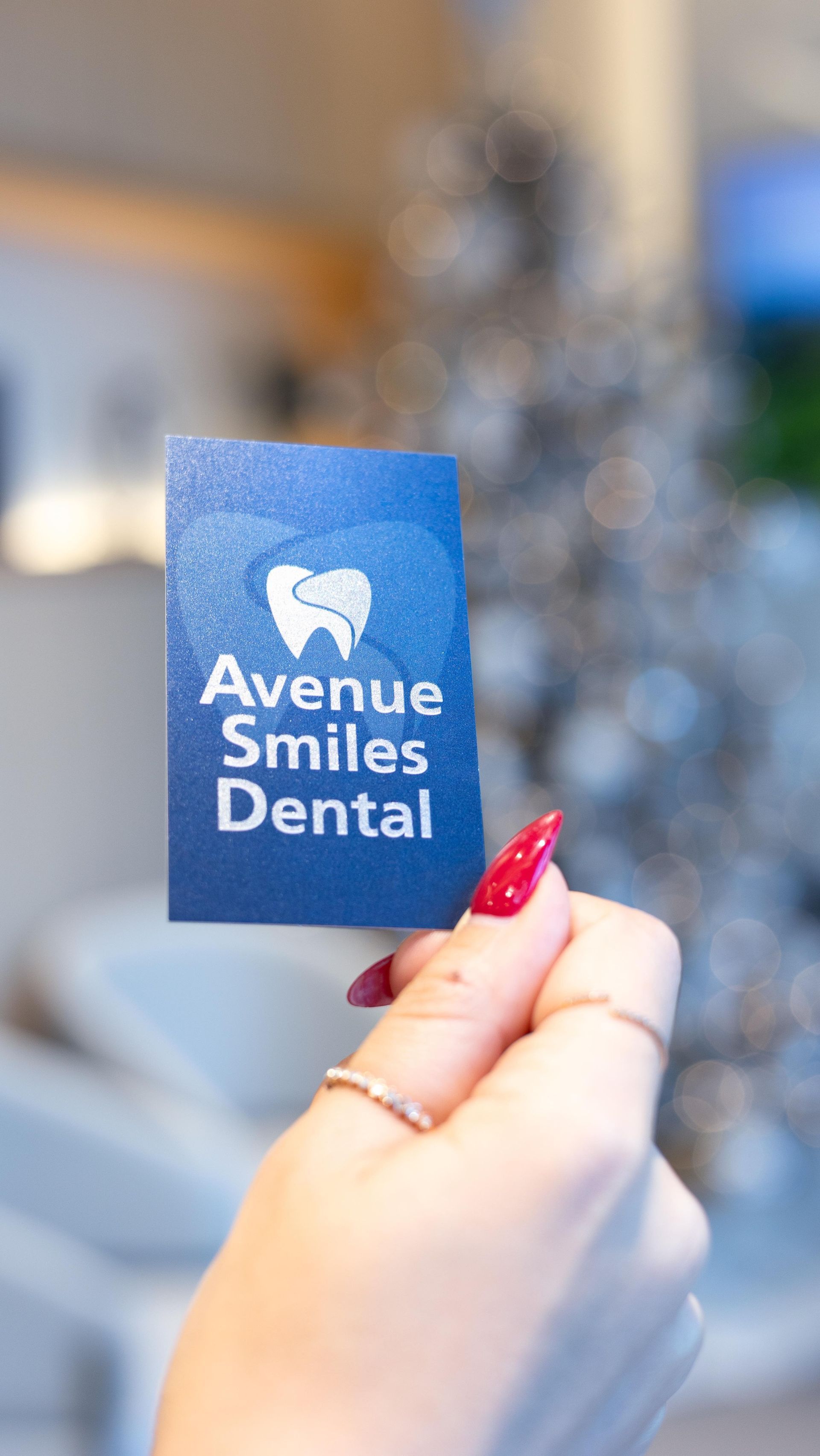 A hand holding a blue Avenue Smiles Dental business card, with a tooth logo, and a glittery background.