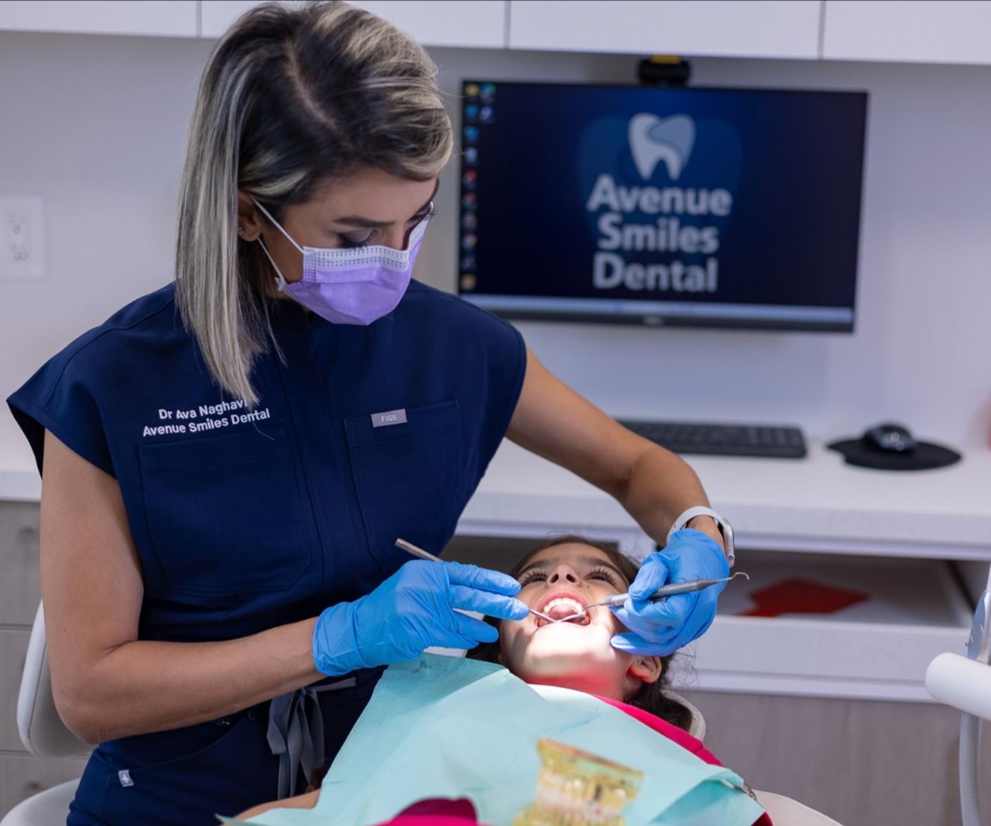 Dentist examines a young girl's teeth at Avenue Smiles Dental, both wearing masks and gloves.