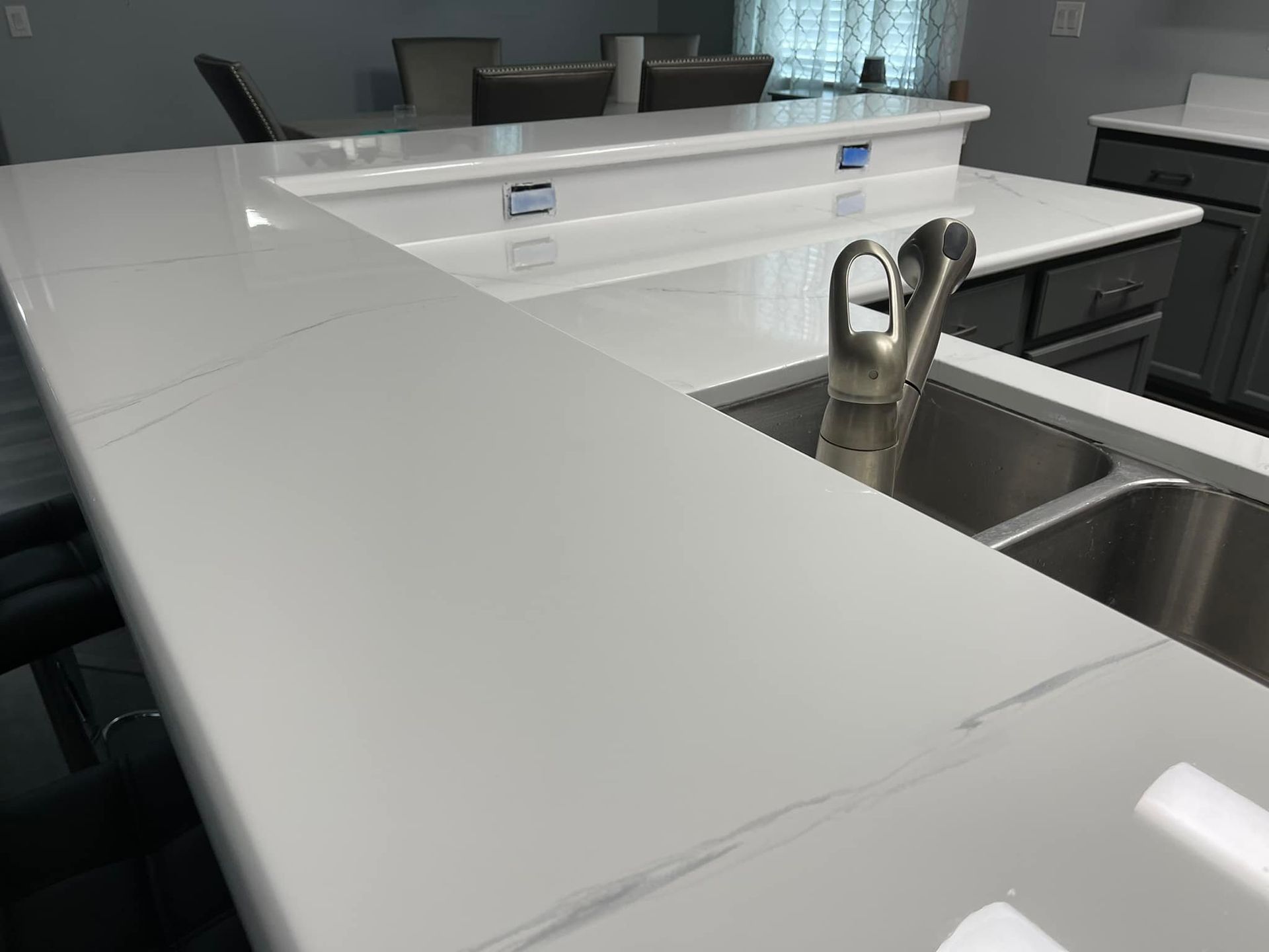 Kitchen island with white countertops, sink, and faucet. Grey cabinets and chairs in the background.