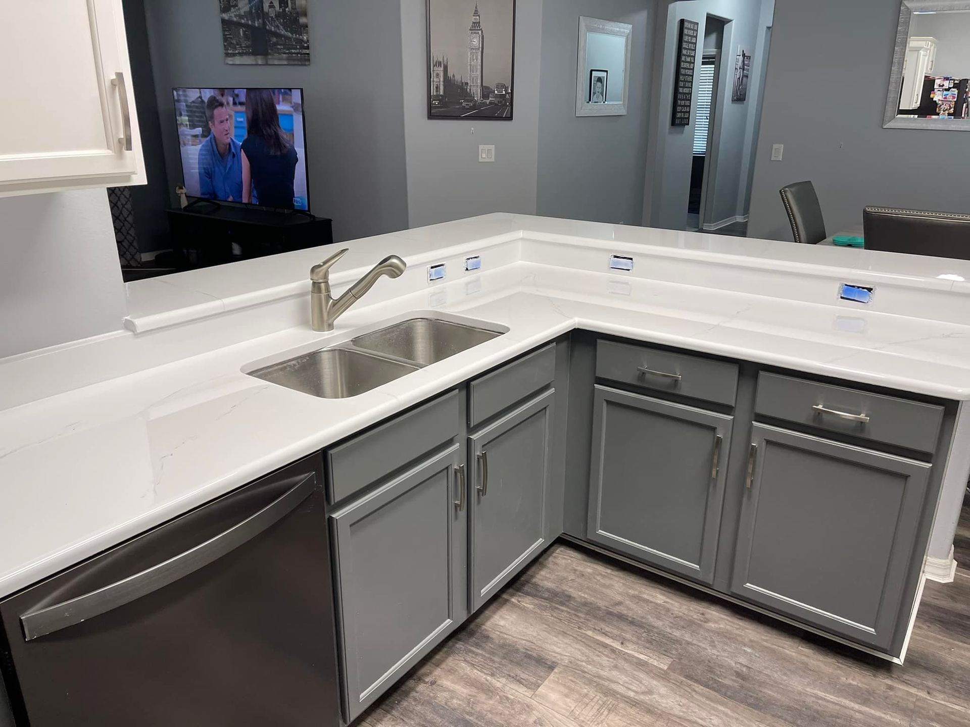 Gray kitchen cabinets with white countertops and a stainless steel sink. A dishwasher is on the left.