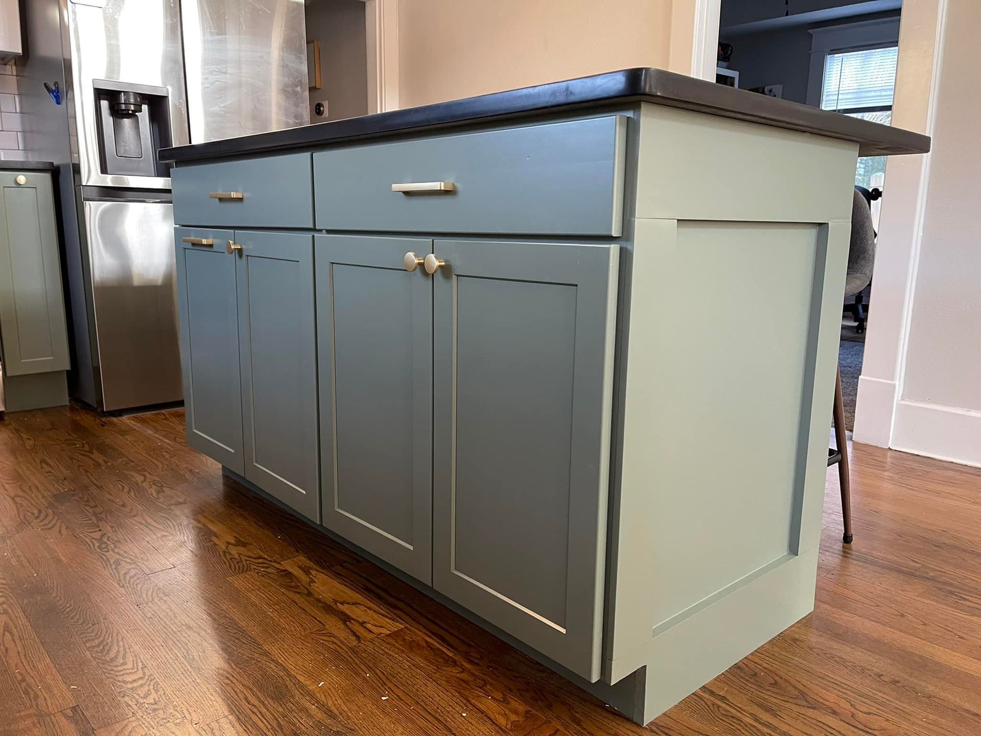 Kitchen island with blue-green cabinets, dark countertop, and gold hardware, on a hardwood floor.
