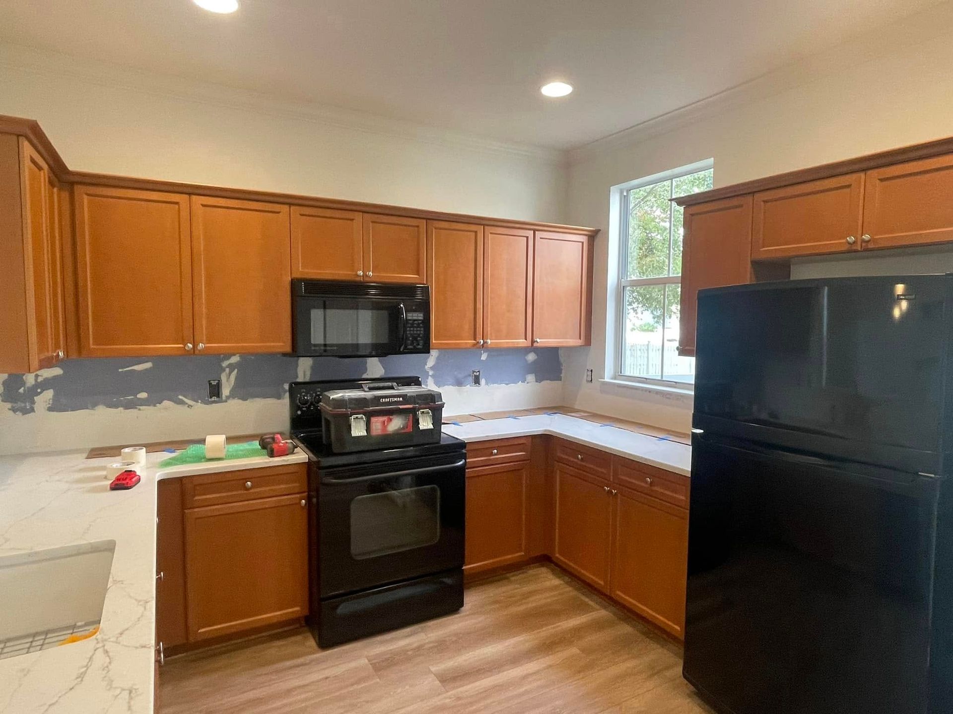 Kitchen with brown cabinets, black appliances, and white countertops. Window in the background.