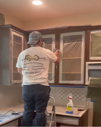 Man spray painting kitchen cabinets; indoor setting with masking.