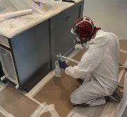Person in protective gear sprays paint on cabinetry in a room.