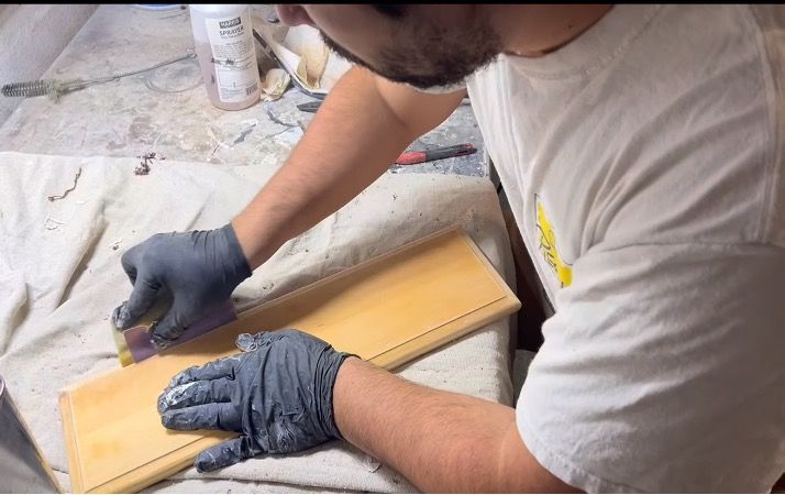 Person wearing gloves sanding a light-colored wooden board on a work surface.