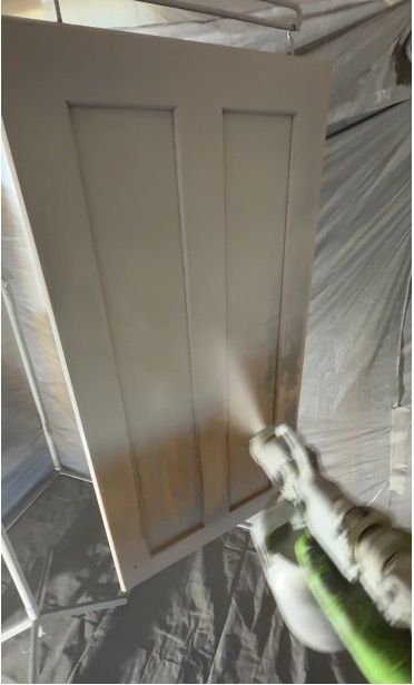 A person spray painting a white cabinet door with a paint sprayer in a workshop.