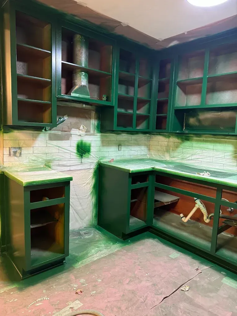 Green painted kitchen cabinets, unfinished, in a room with a light fixture, paint splatters on backsplash and countertop.