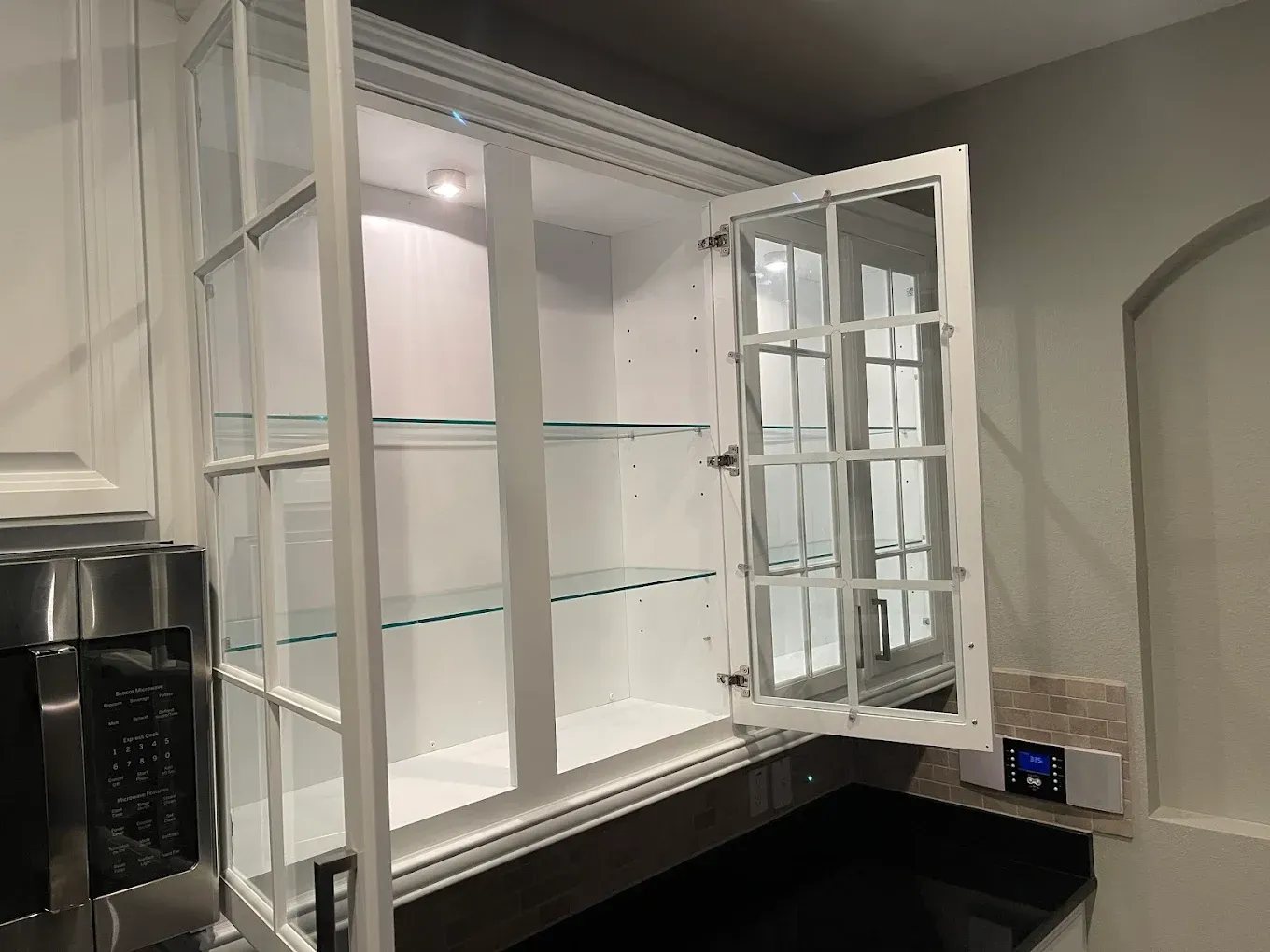 White kitchen cabinet with glass doors, one open, above dark countertop.