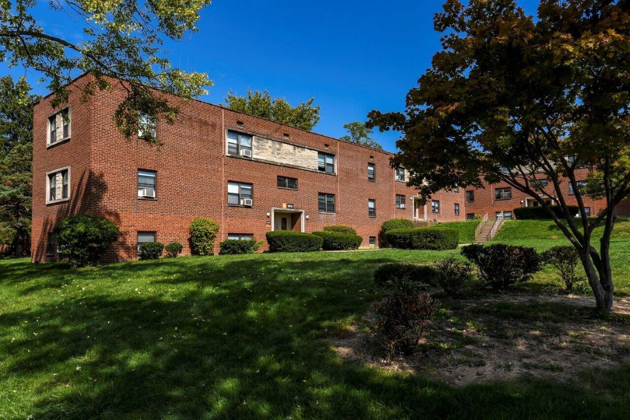 Red brick apartment building on a grassy hill under a blue sky, trees in the foreground.