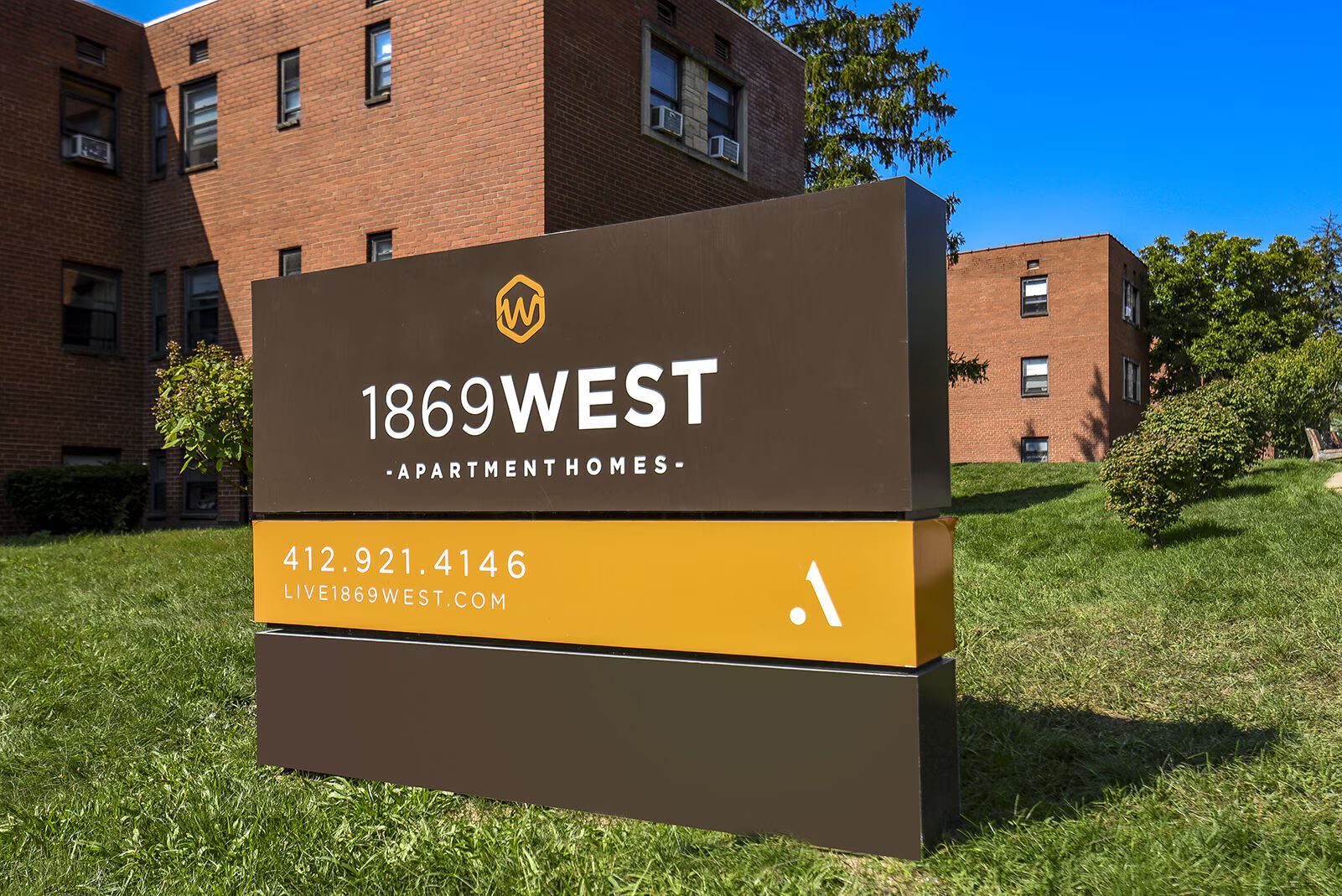 Sign for 1869 West Apartments; brown and gold with address and phone number on green lawn, brick building background.
