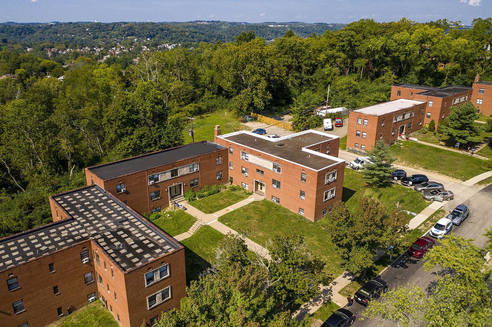 Brick apartment buildings surrounded by green trees on a hillside with parked cars.
