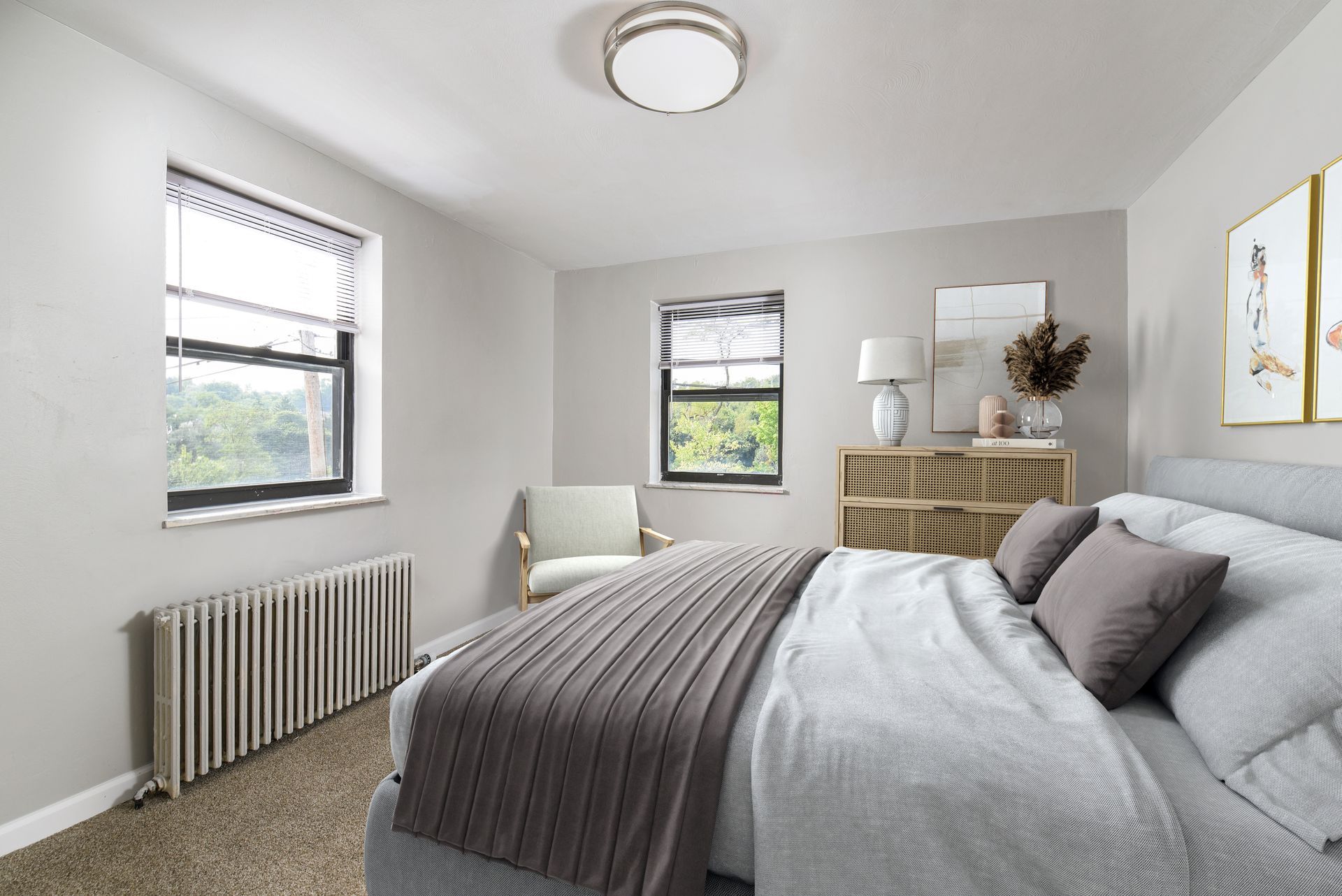 Bedroom with bed, dresser, chair, two windows, and radiator. Neutral colors and natural light.