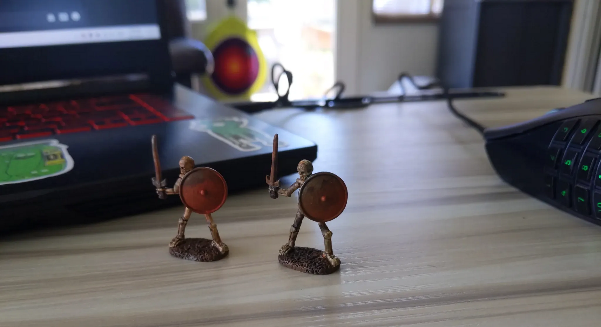 Two miniature toy warriors with swords and shields stand on a desk near a laptop and mouse.