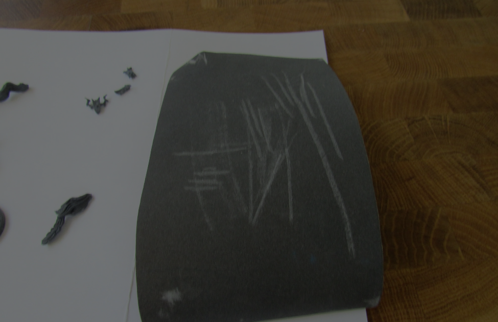 A large, dark package in clear plastic wrap sits on white paper; small, dark objects lie scattered nearby.
