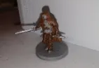 Miniature figure of a warrior with two swords, brown and white armor, on a gray base.
