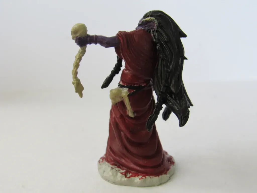 Miniature figure of a humanoid in a red robe, holding a skull and long braid; dark hair and purple skin.