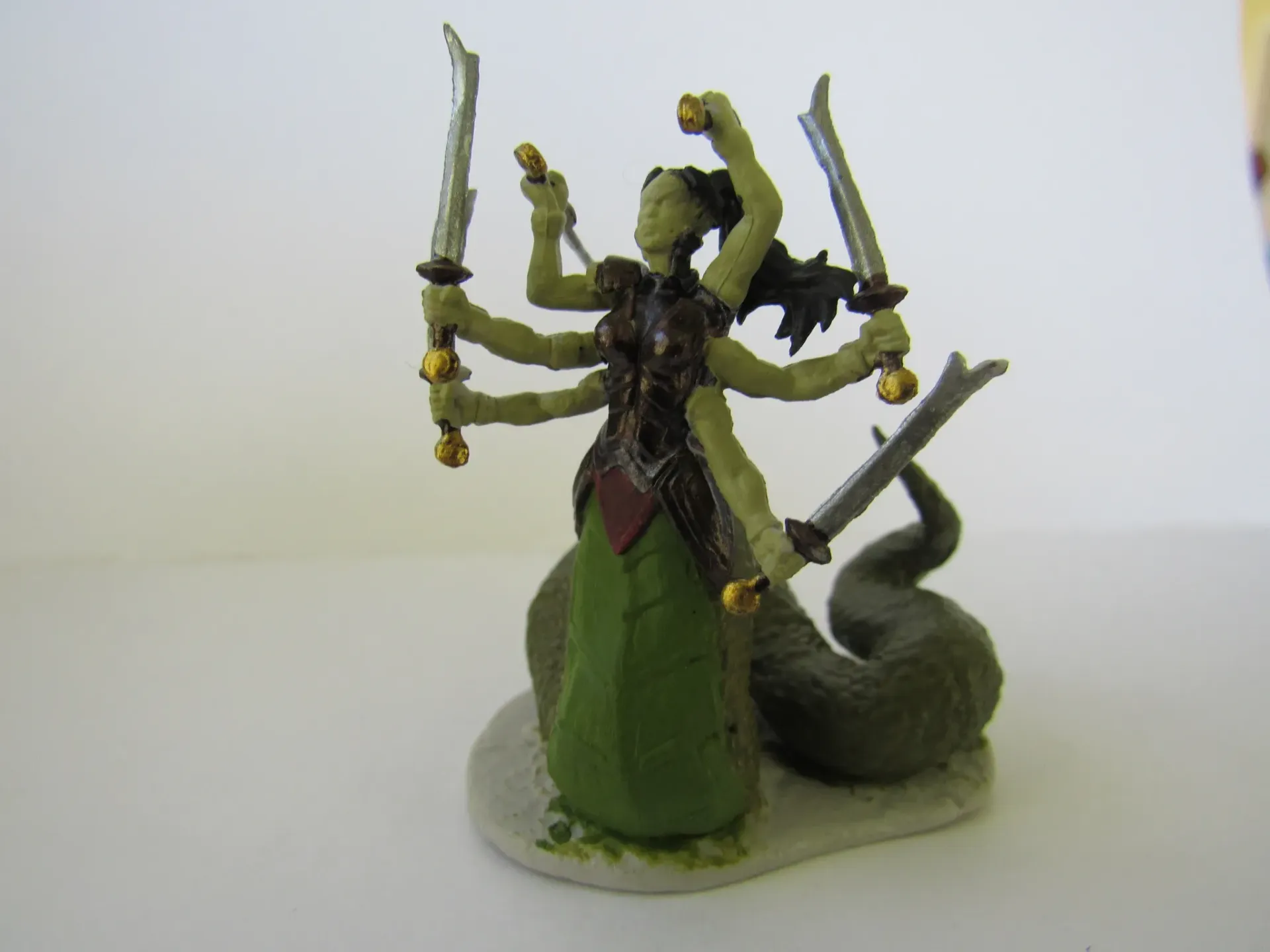 A green-skinned, six-armed snake-bodied figure holding swords, wearing a dark vest, on a white base.
