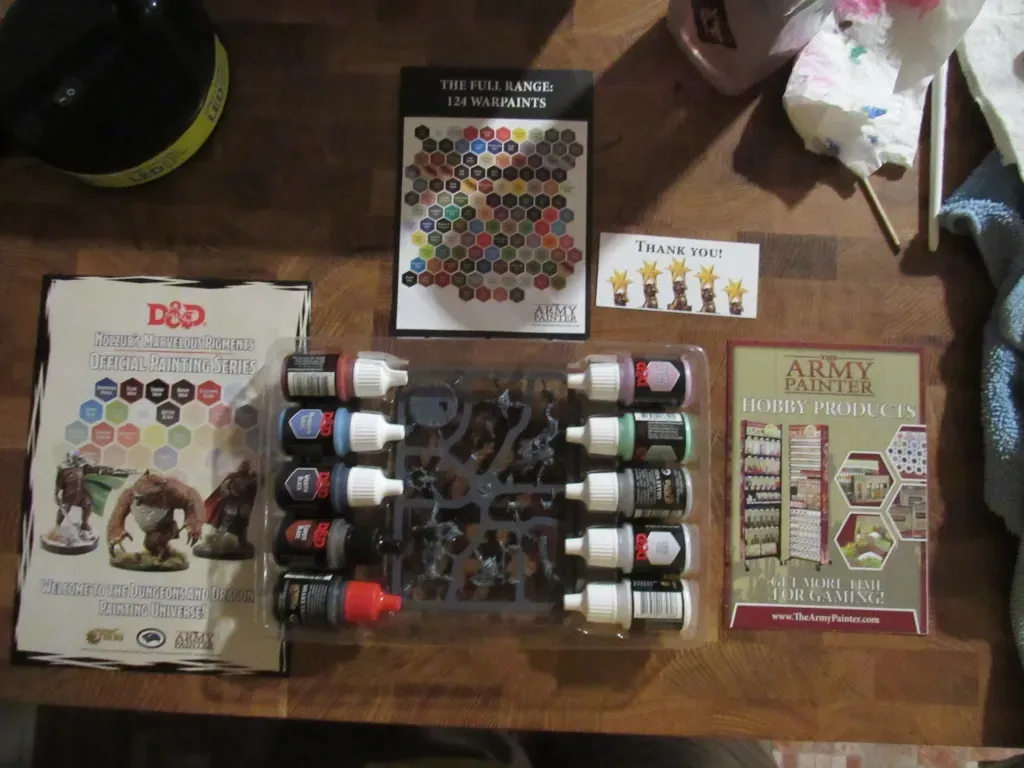 Miniature painting kit with paints, figures, stickers, and instruction cards on a wooden table.