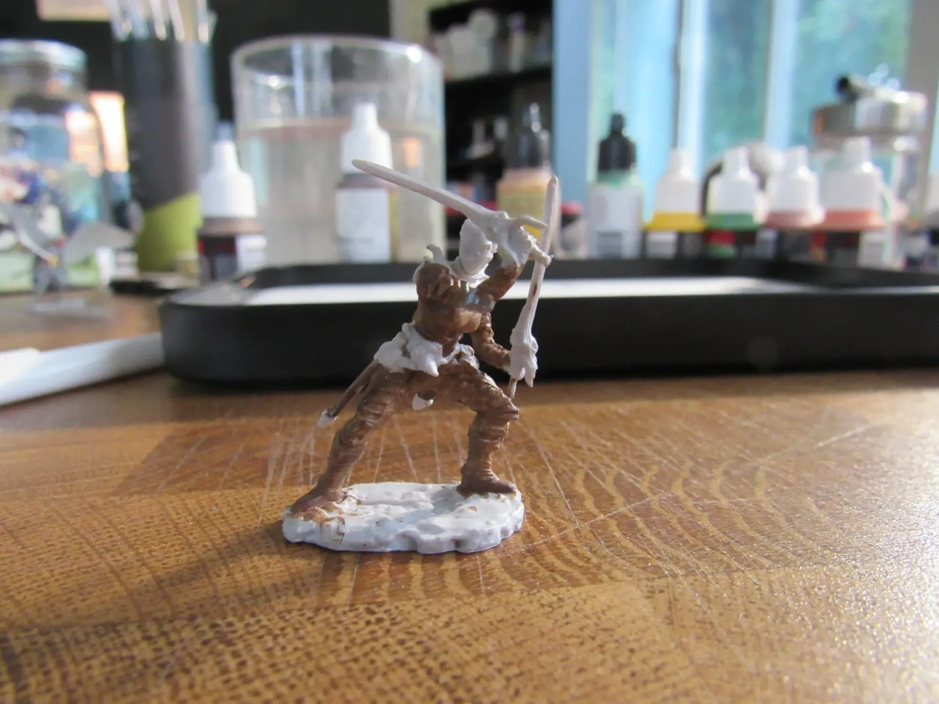 Miniature figure of a warrior painted brown and white on a wooden surface with painting supplies in the background.