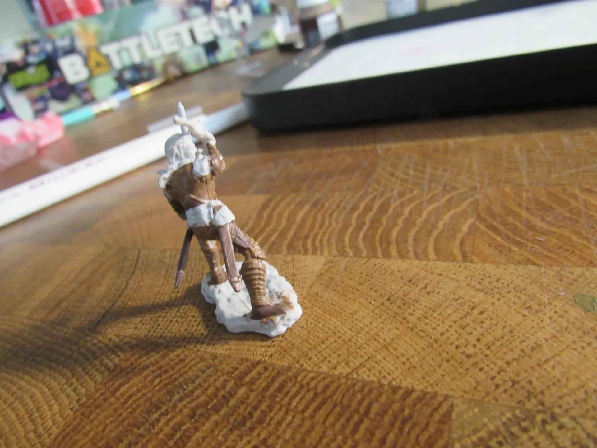 Miniature figure on wooden surface, partially painted in brown and tan, with white base.