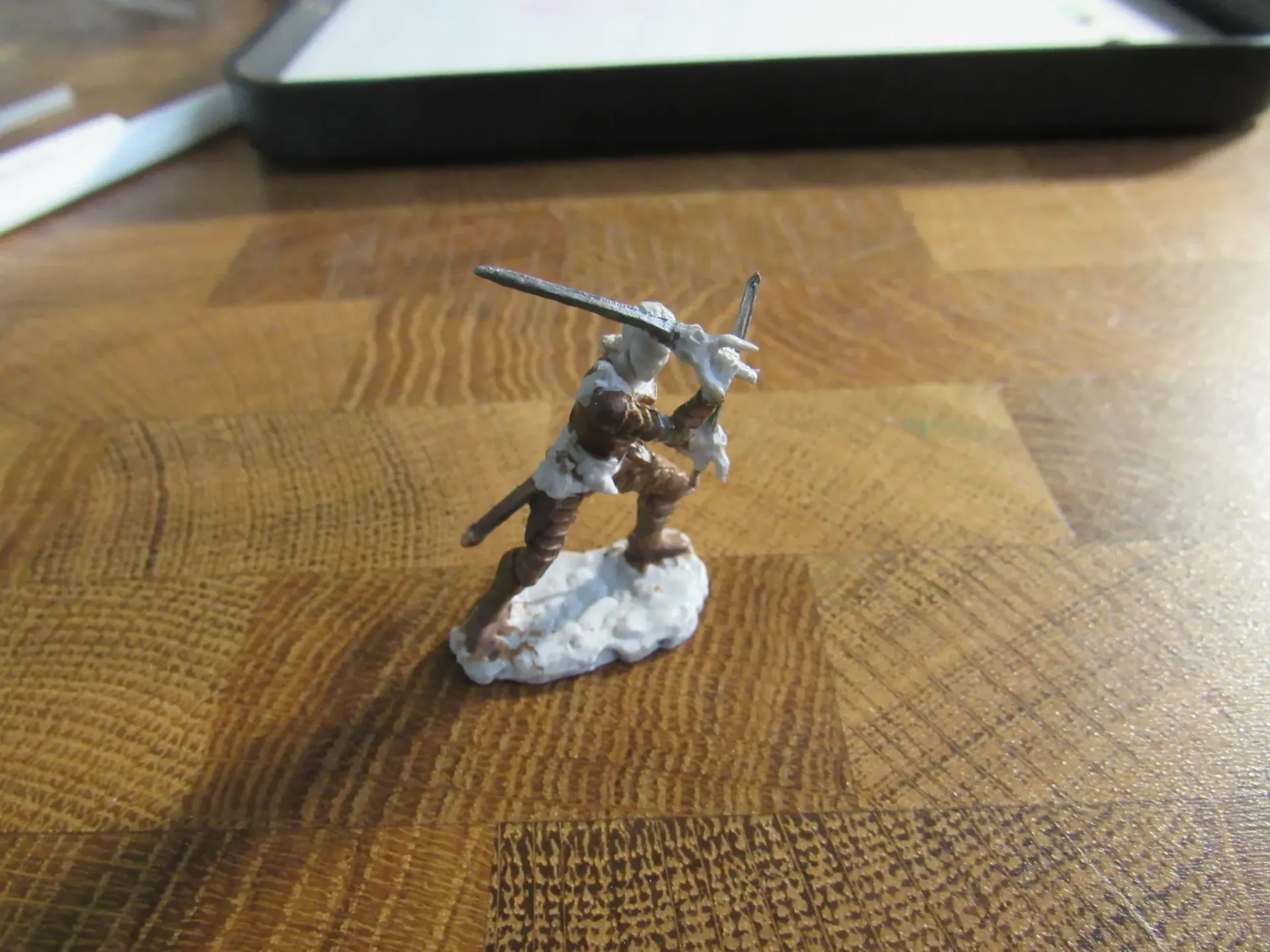 Miniature figure of a person with a sword in a battle stance, on a textured wooden surface.