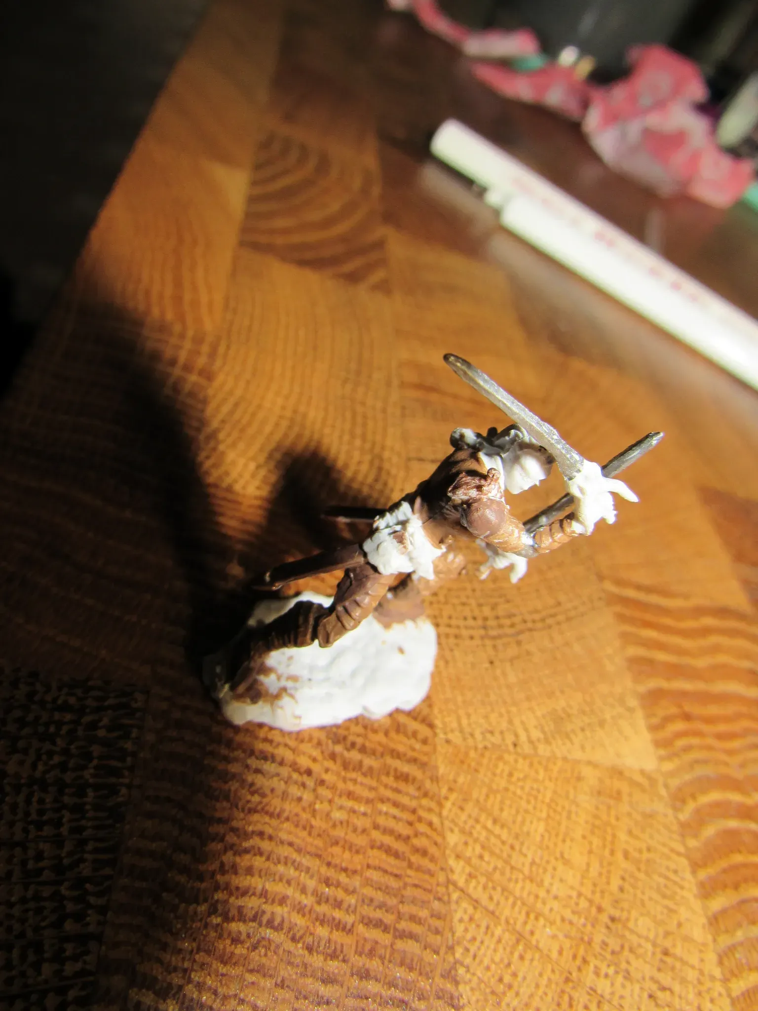 Miniature figure of a brown creature with white accents, holding two swords, on a wooden surface.