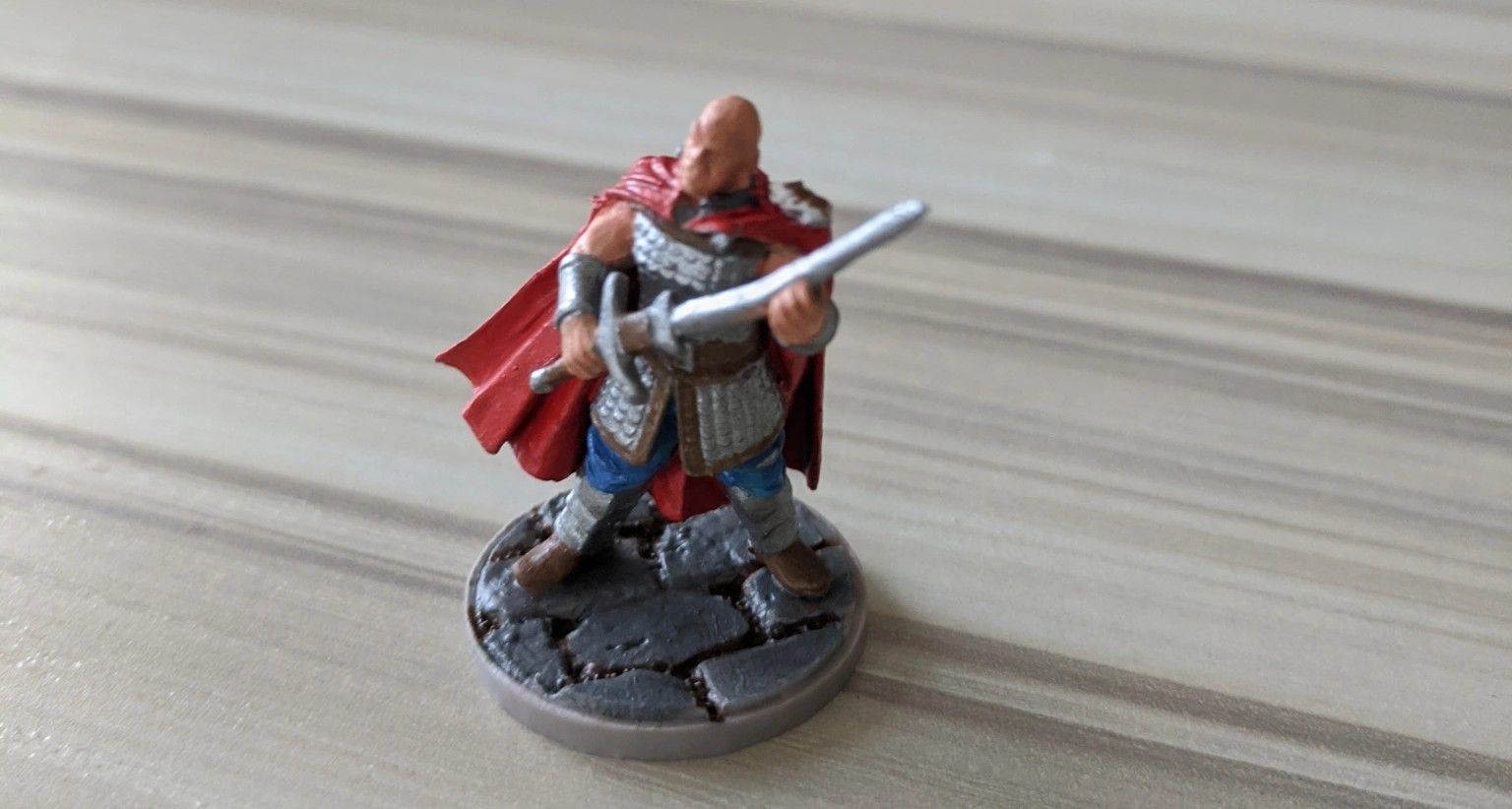 Miniature of a bald warrior with a red cape holding a sword, standing on a stone base.