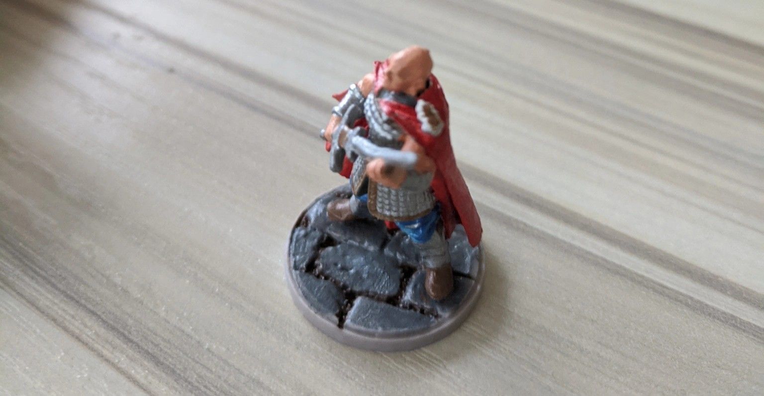 Miniature figure of a bald warrior in red cape, armor, and sword on a cobblestone base.