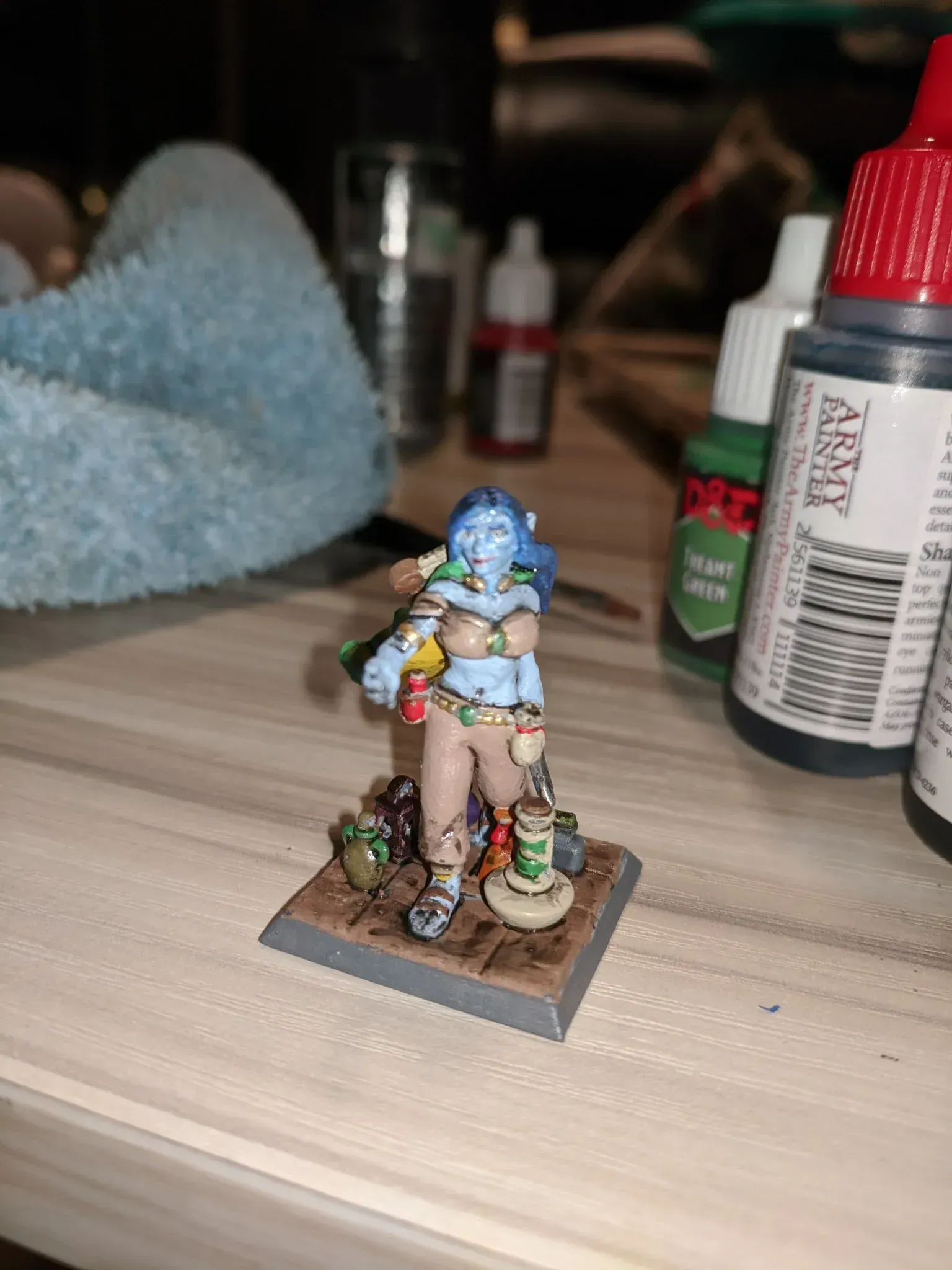 Miniature figure of a blue-skinned woman, holding a wand, on a square base. She's on a table, with paint bottles in the background.