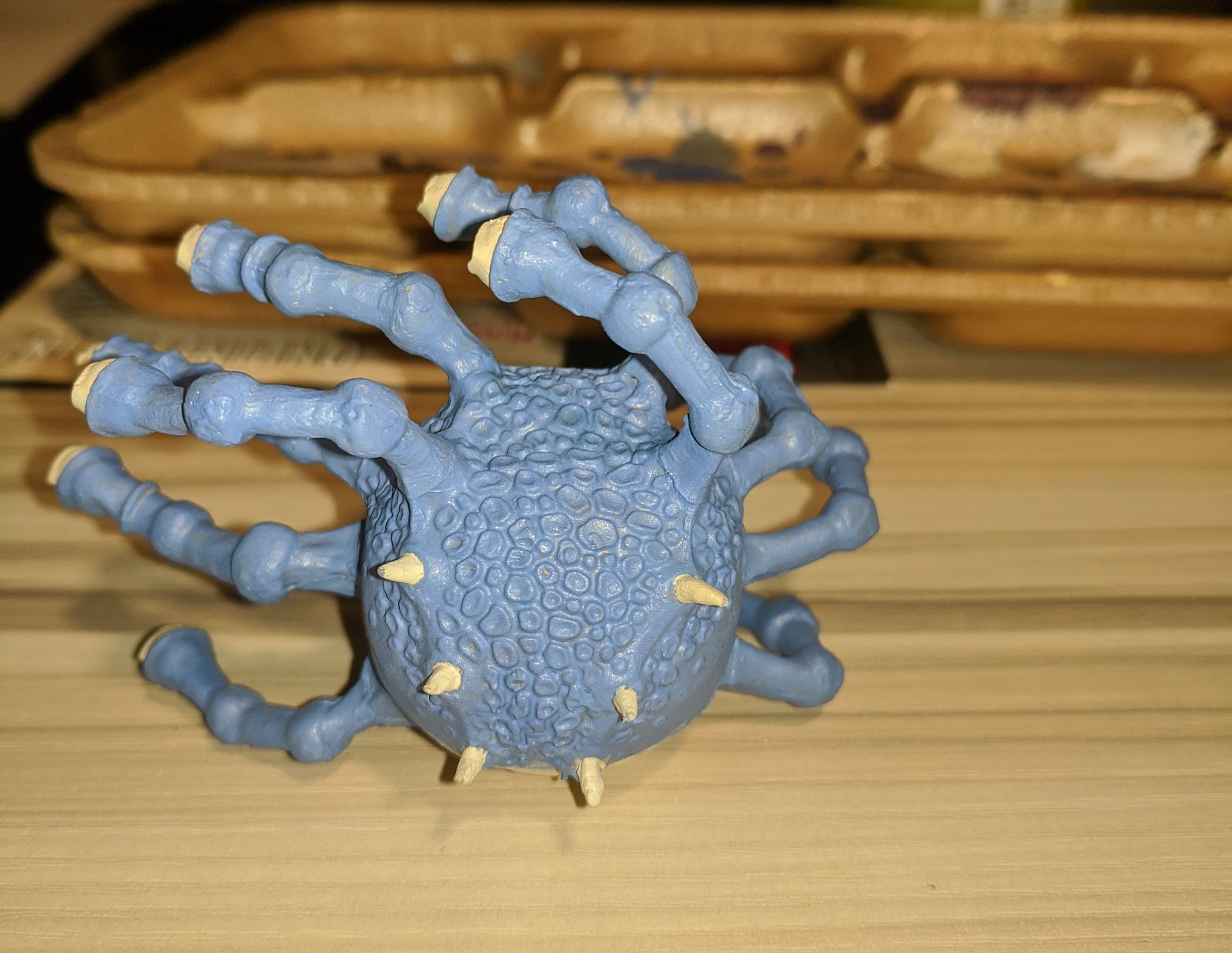 A blue, spiky ball-shaped model with arms and yellow tips sits on a wooden surface.