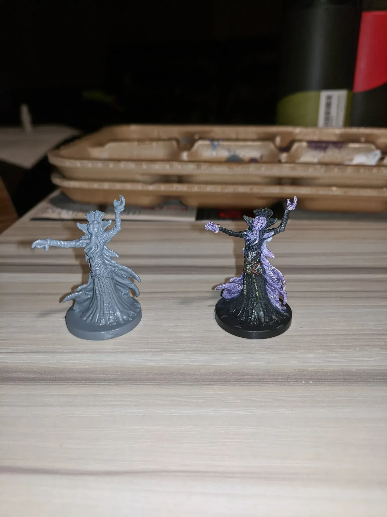 Two miniature figures, one unpainted gray, the other painted black, green and purple, arms outstretched.