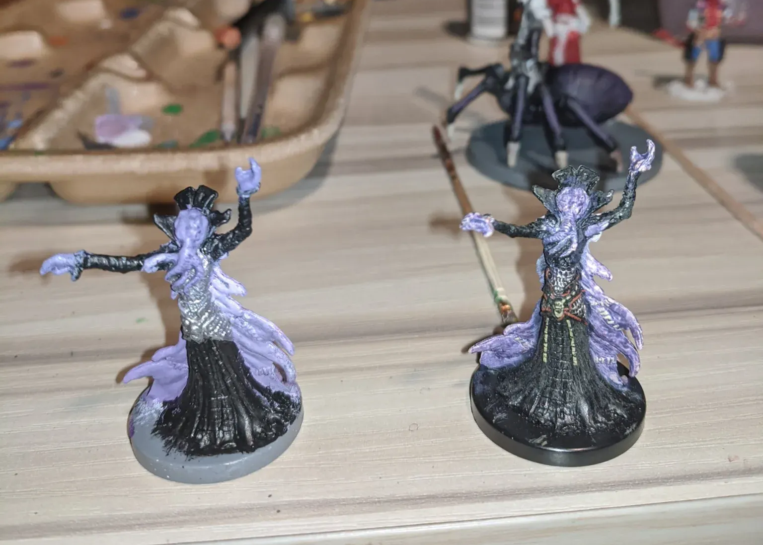 Two painted miniatures of fantasy figures with flowing purple and black robes and arms outstretched on a table.