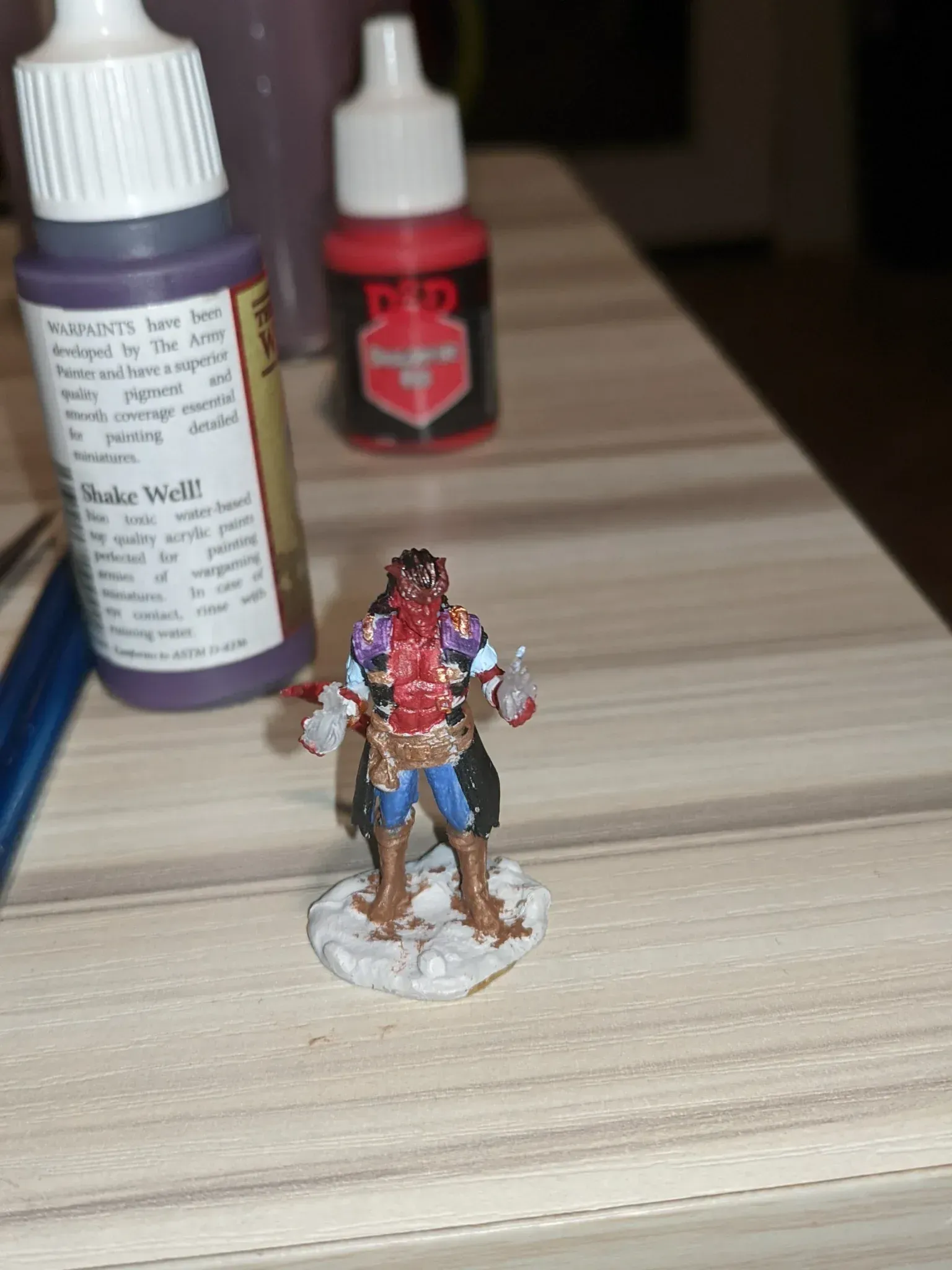 Miniature tabletop figure, painted with blue pants, red torso, standing on a white base, next to paint bottles.