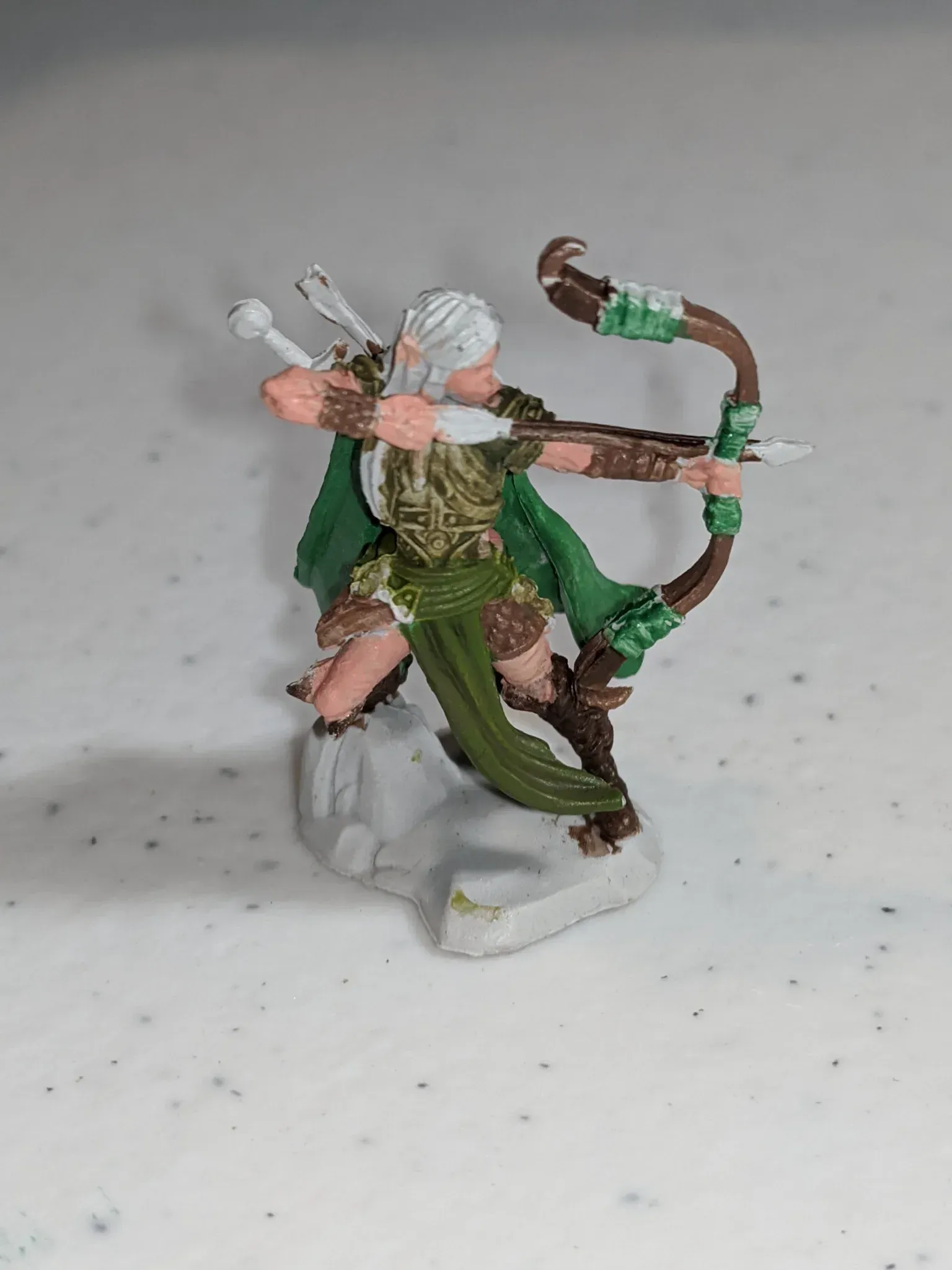 Elf archer figurine, white hair, green cloak and bow, brown and gold armor, aiming at a target.