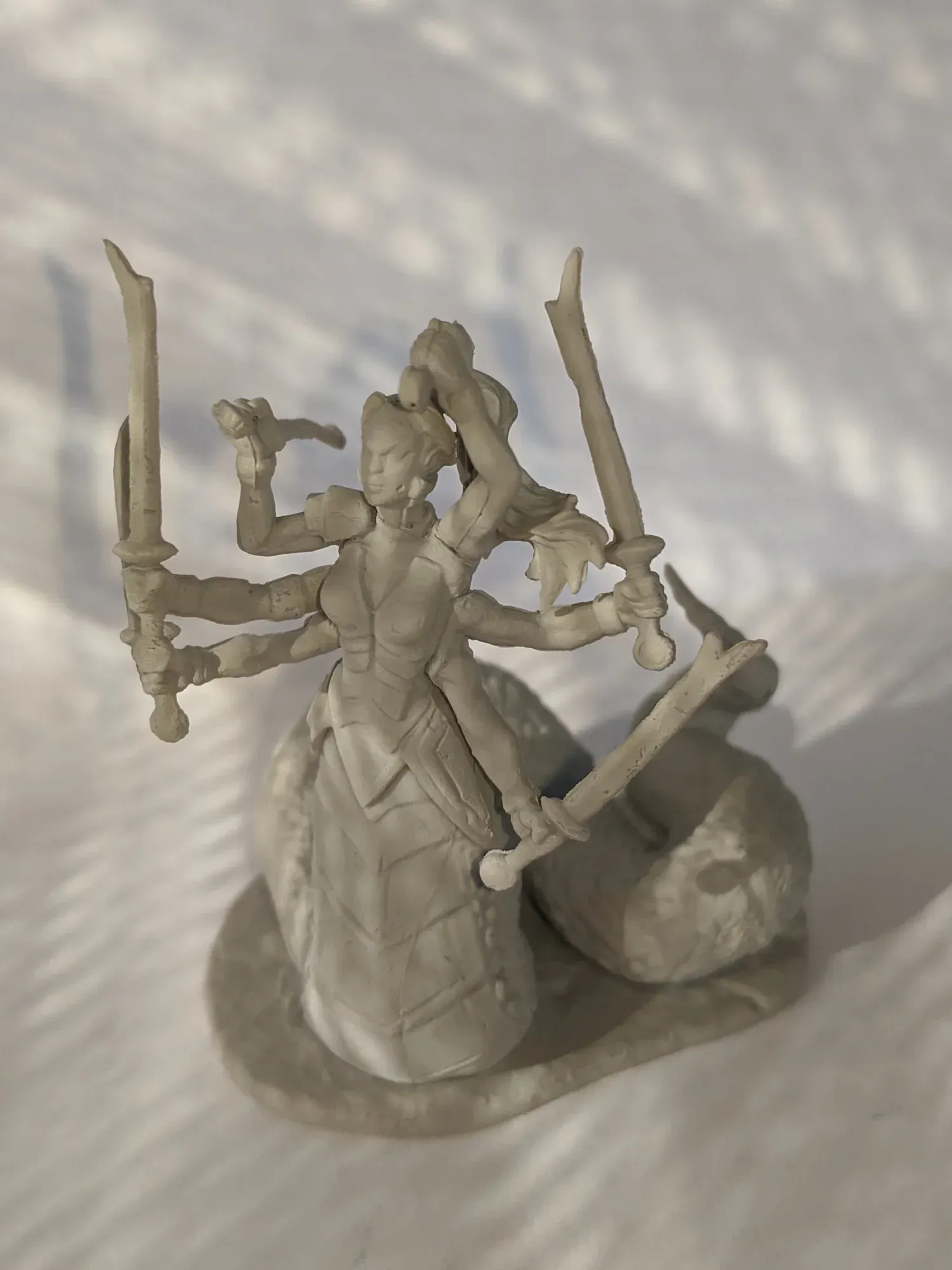 Gray miniature figure of a woman with six arms wielding swords, atop a snake-like body.