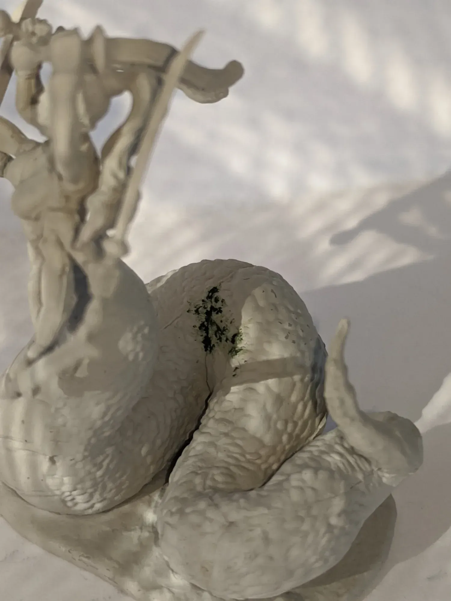 A light-colored sculpted snake figure with a dark green spot on its back; sunny setting.
